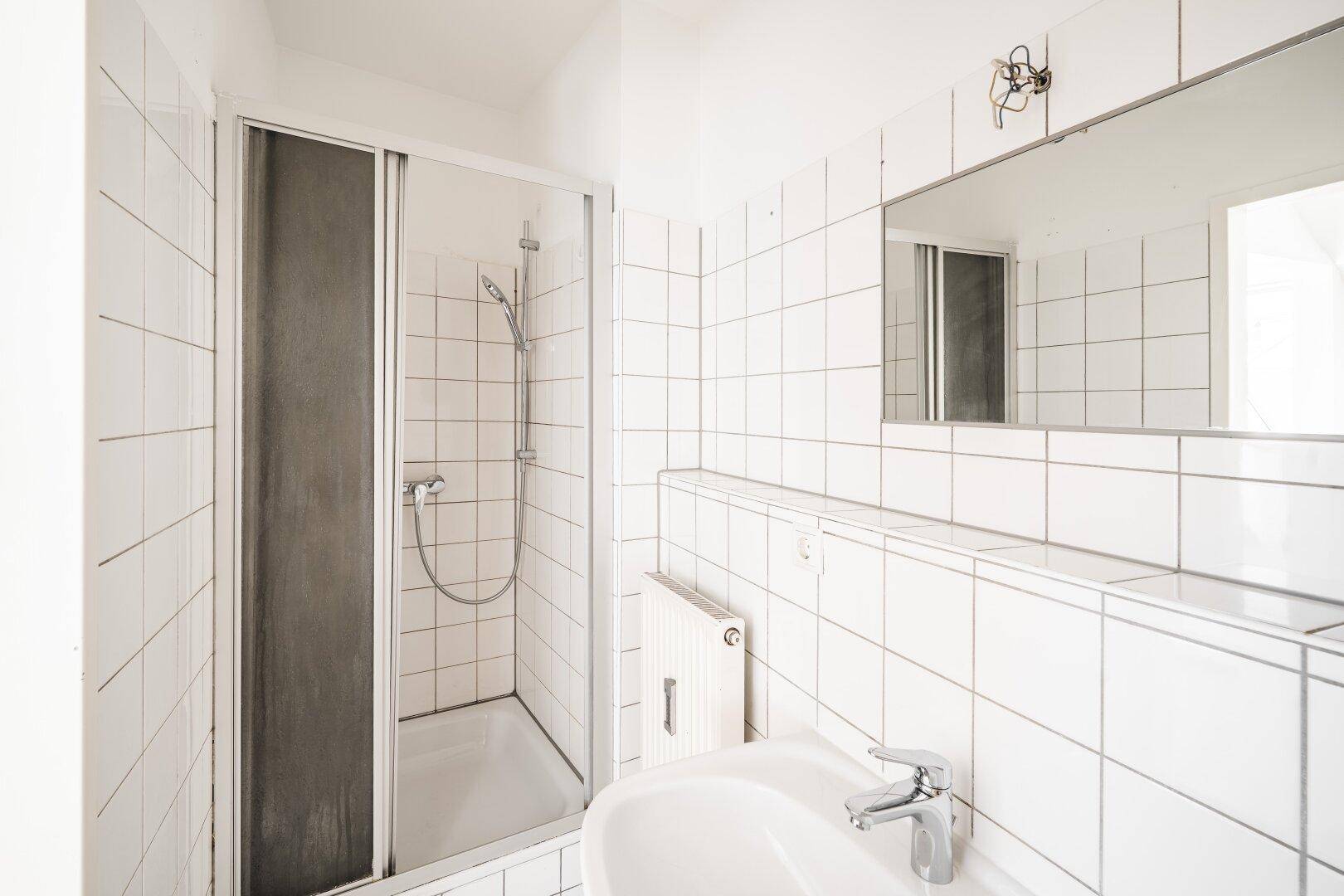 Funktionales Badezimmer mit Duschkabine, Waschbecken und weißen Fliesen, ausgestattet mit einem Heizkörper.