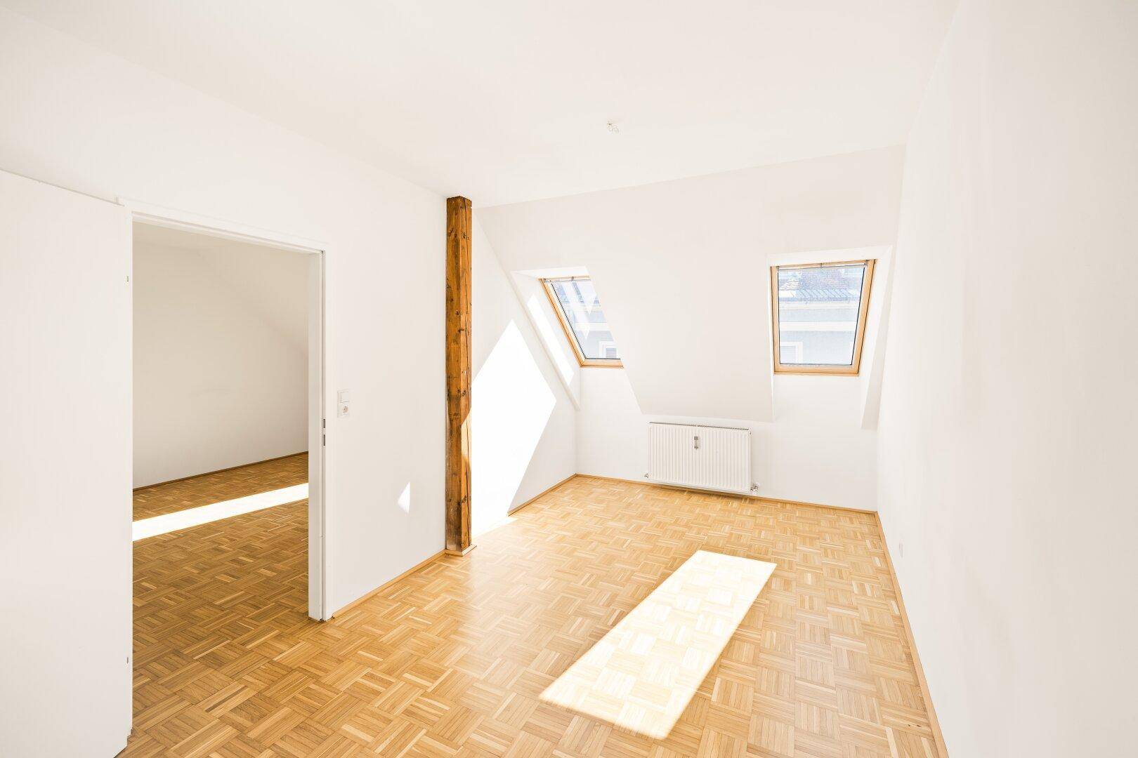 Helles Zimmer mit Dachschrägen und zwei Dachfenstern, ausgestattet mit Parkettboden und einem Holzbalken.