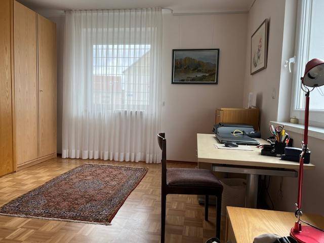 Schlafzimmer mit Holzschrank, Schreibtisch und hellem Fenster mit weißen Vorhängen.