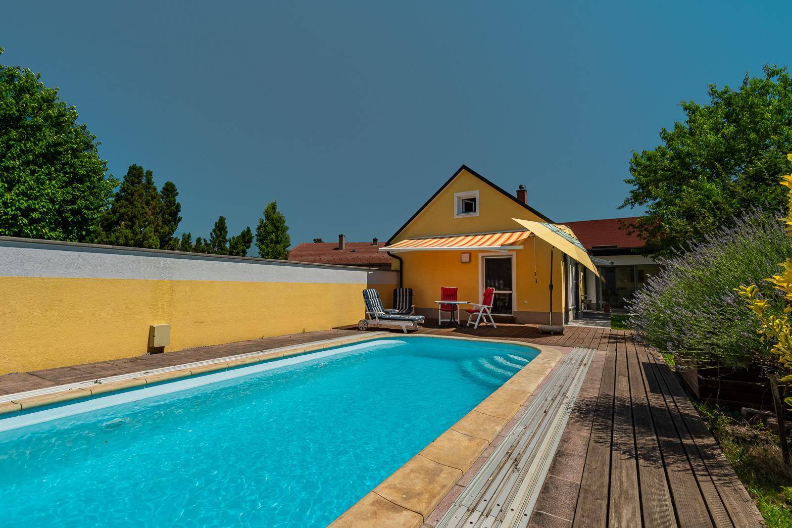 Außenbereich mit großem Pool, Holzdeck und Liegestühlen unter blauem Himmel am Haus.