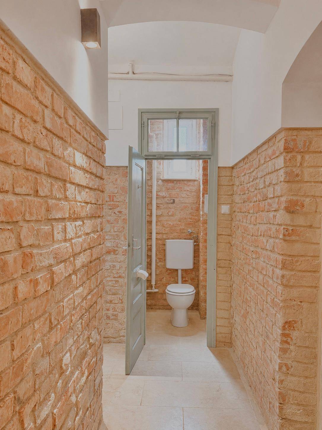 Schmaler Flur mit unverputzten Ziegelwänden und Blick auf eine Toilette.
