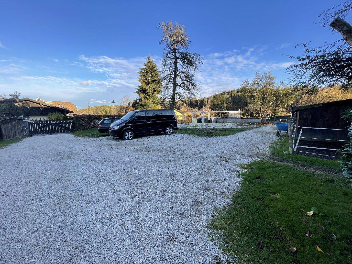Geräumiger Schotterparkplatz mit abgestellten Fahrzeugen, umgeben von Bäumen und Gebäuden.