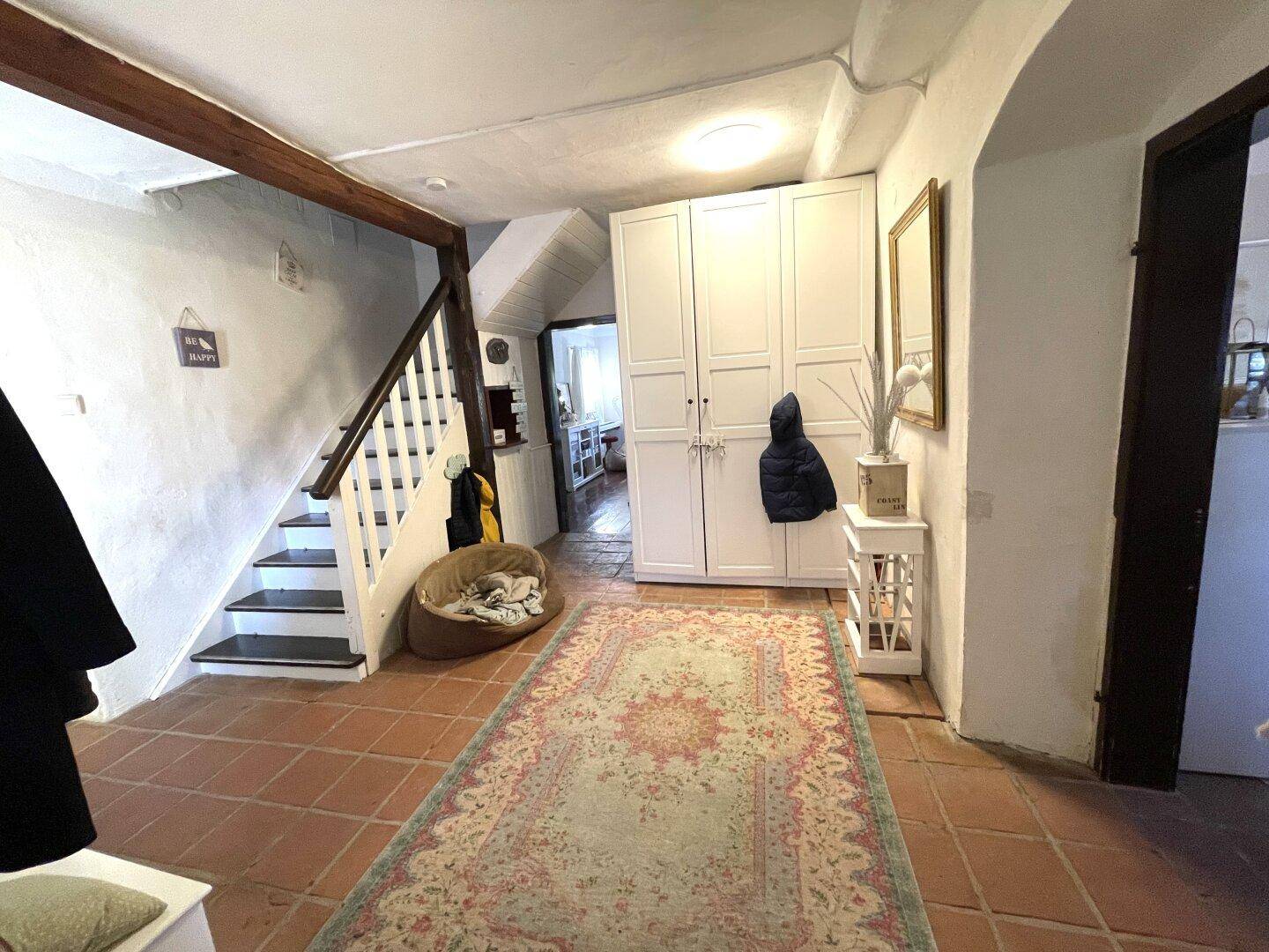 Eingangsbereich mit Terrakottaboden, Holztreppe und weißem Schrank.