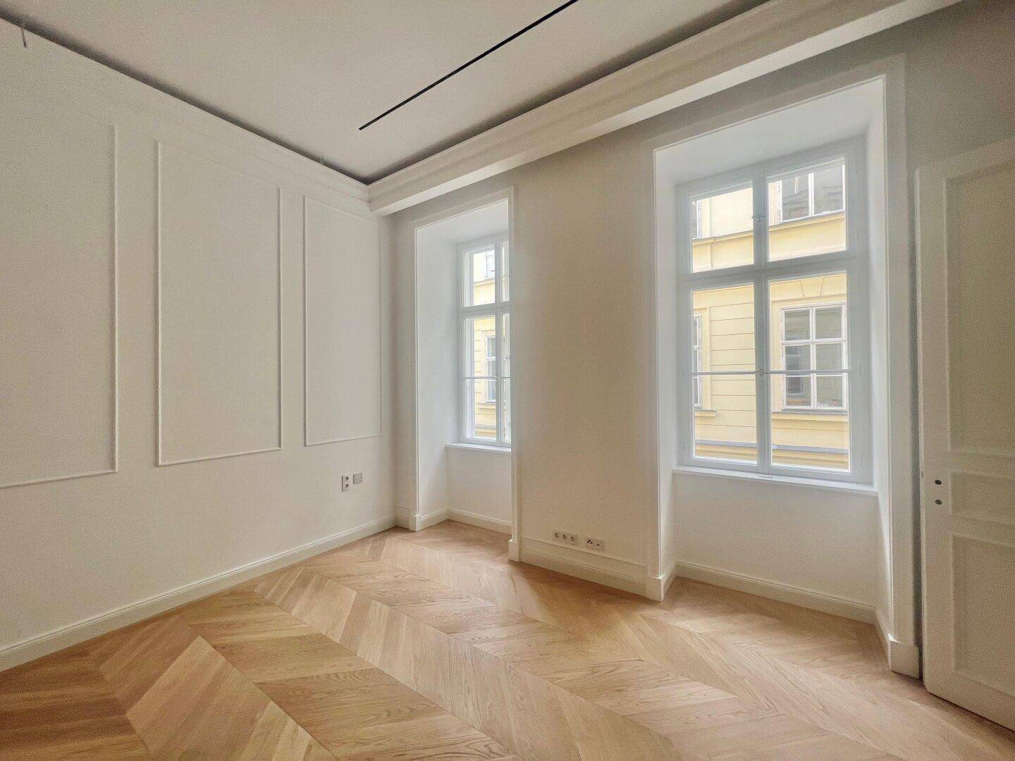 Lichtdurchfluteter Raum mit Fischgrätparkett, hohen Fenstern und klassischer Wandgestaltung.
