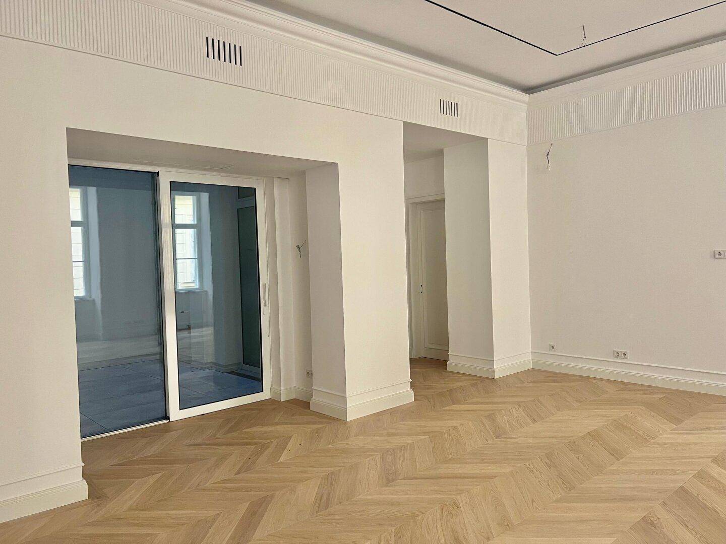 Großer, heller Raum mit Fischgrätparkett und Schiebetür zu einem angrenzenden Bereich.