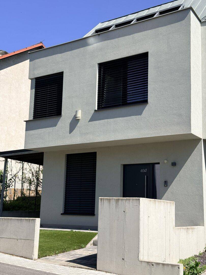 Moderne Hausfassade mit großen Fenstern, Rollläden und gepflegtem Vorgarten.