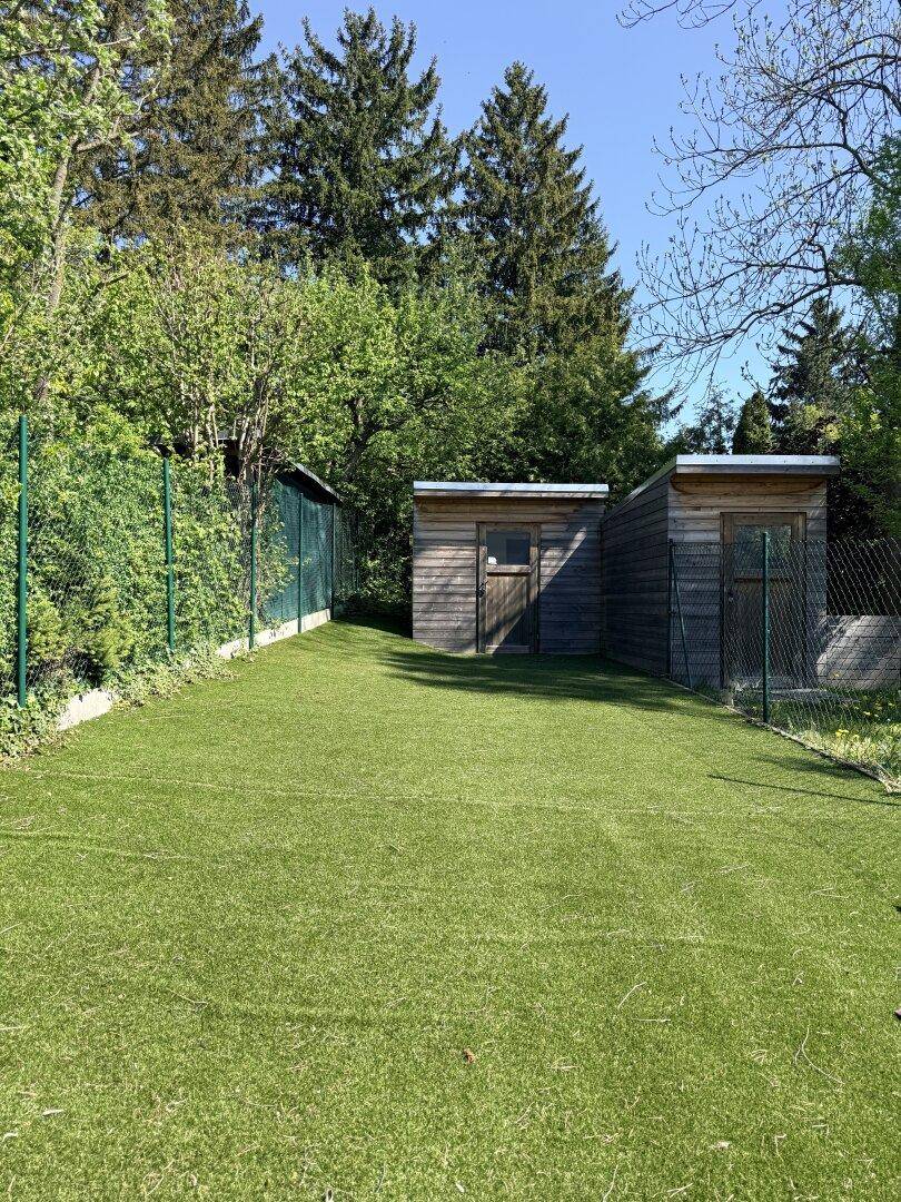 Großer Gartenbereich mit Kunstrasen und zwei Holzschuppen umgeben von Bäumen.