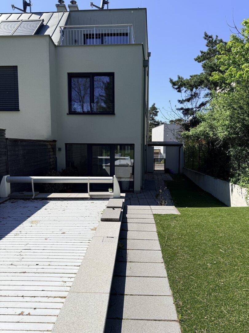 Gartenbereich mit Holzterrasse, Rasenfläche und modernem Gebäude im Hintergrund.