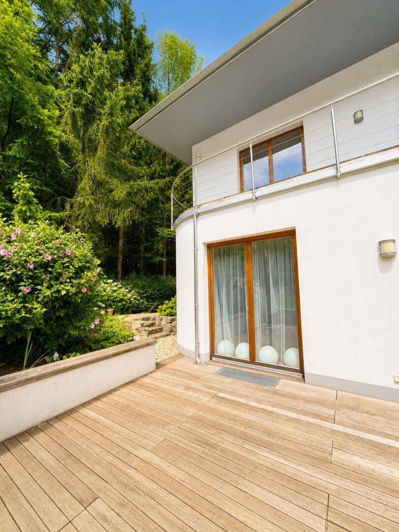 Moderne Hausfassade mit geschwungener Architektur und Holzterrasse umgeben von viel Grün.