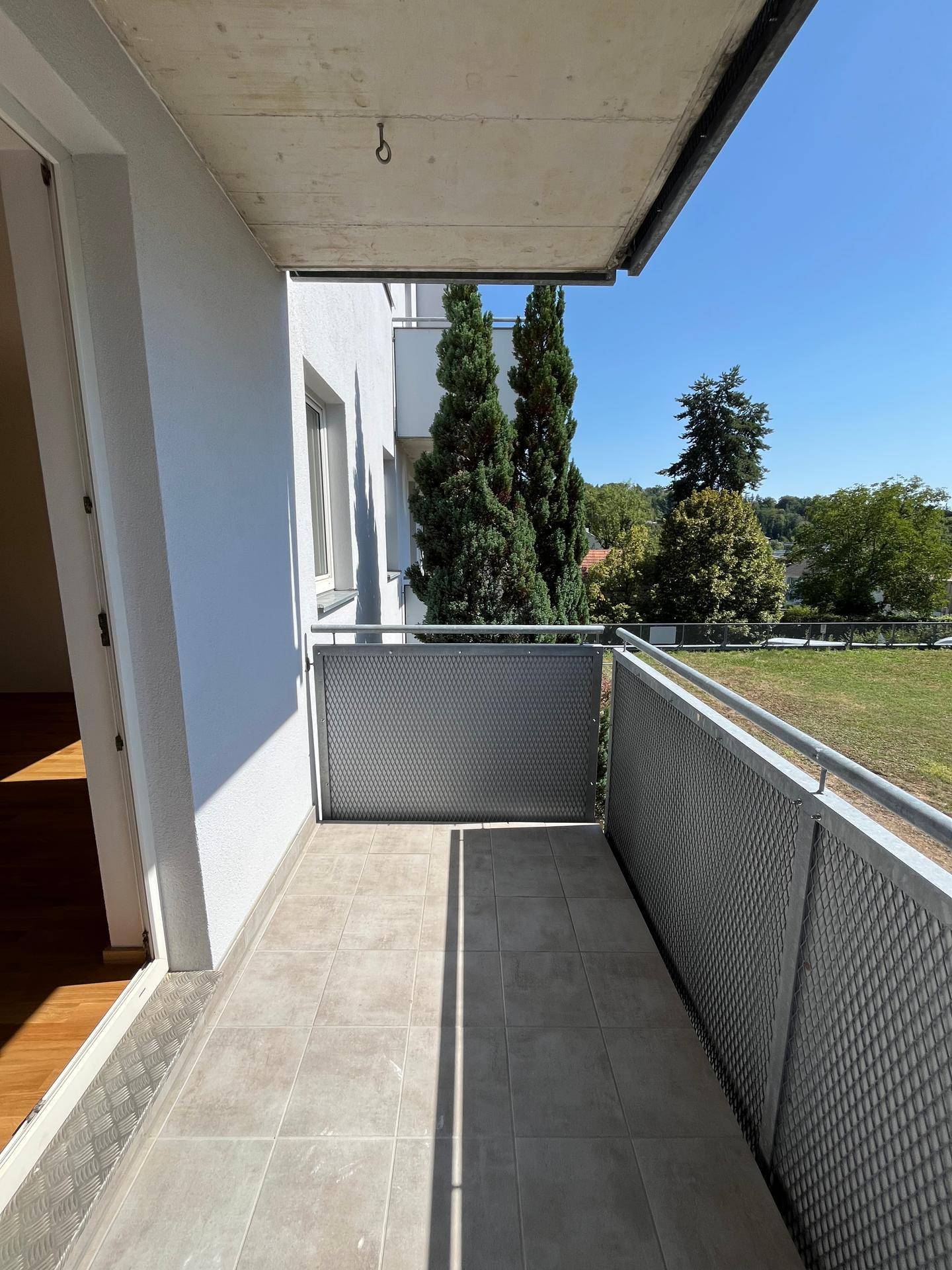 Schmaler Balkon mit Fliesenboden und Metallgeländer, bietet eine Aussicht auf Bäume und Rasenflächen.