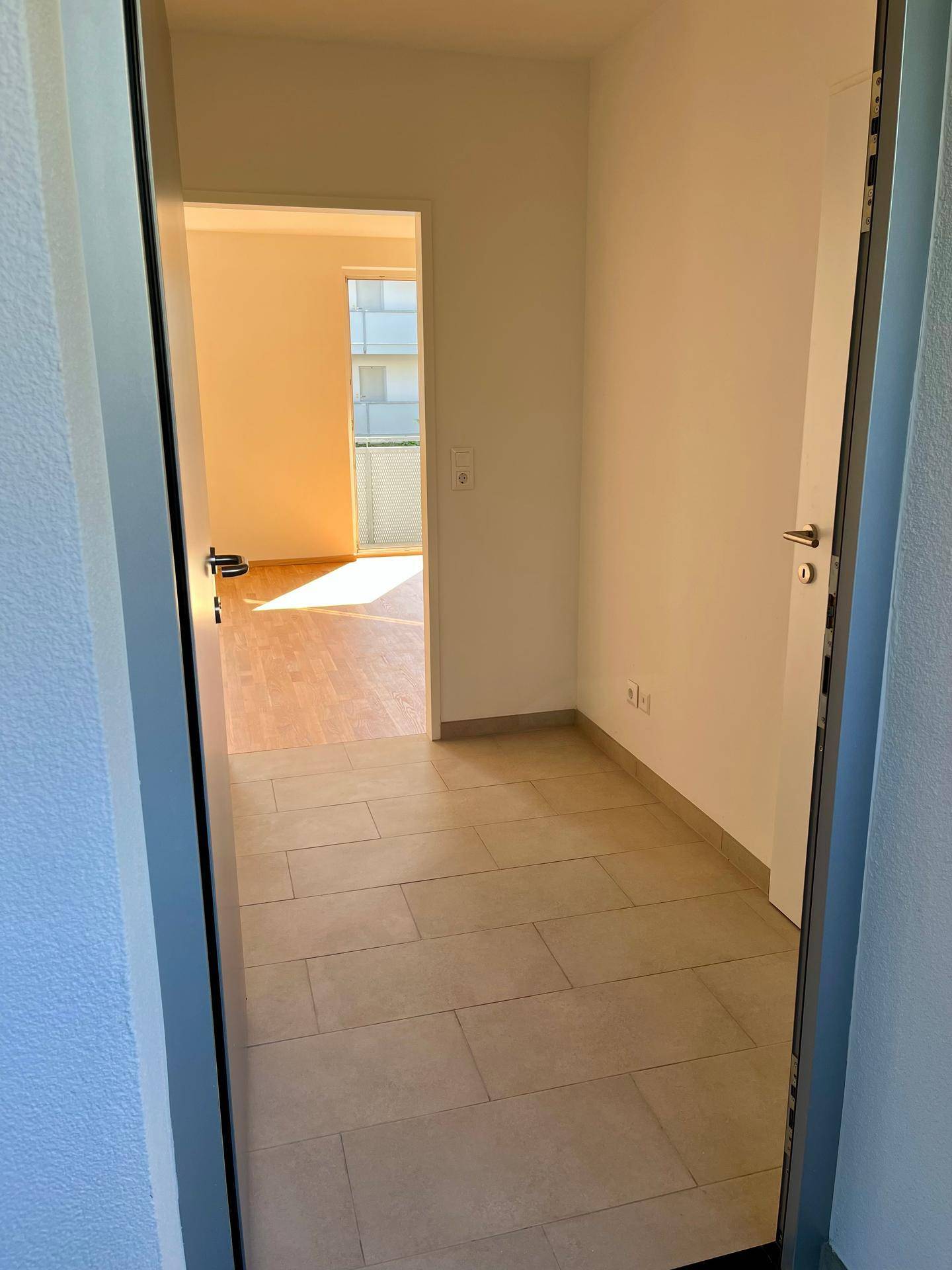 Eingangsbereich mit Fliesenboden, der in ein helles Zimmer mit Holzboden und Balkonzugang führt.