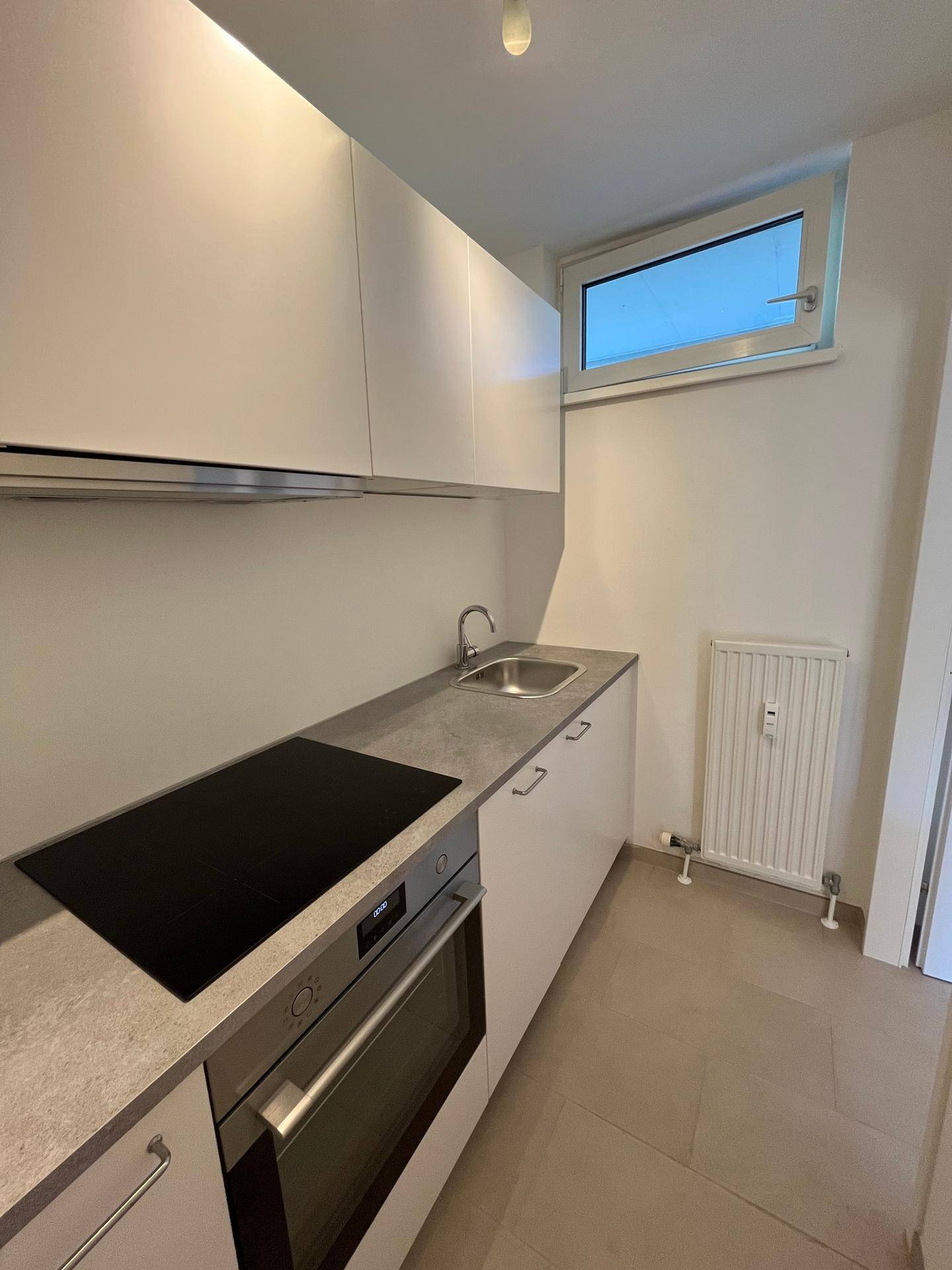 Helle Küche mit Fenster, weißen Schränken, Spüle und integriertem Kochfeld und Backofen.