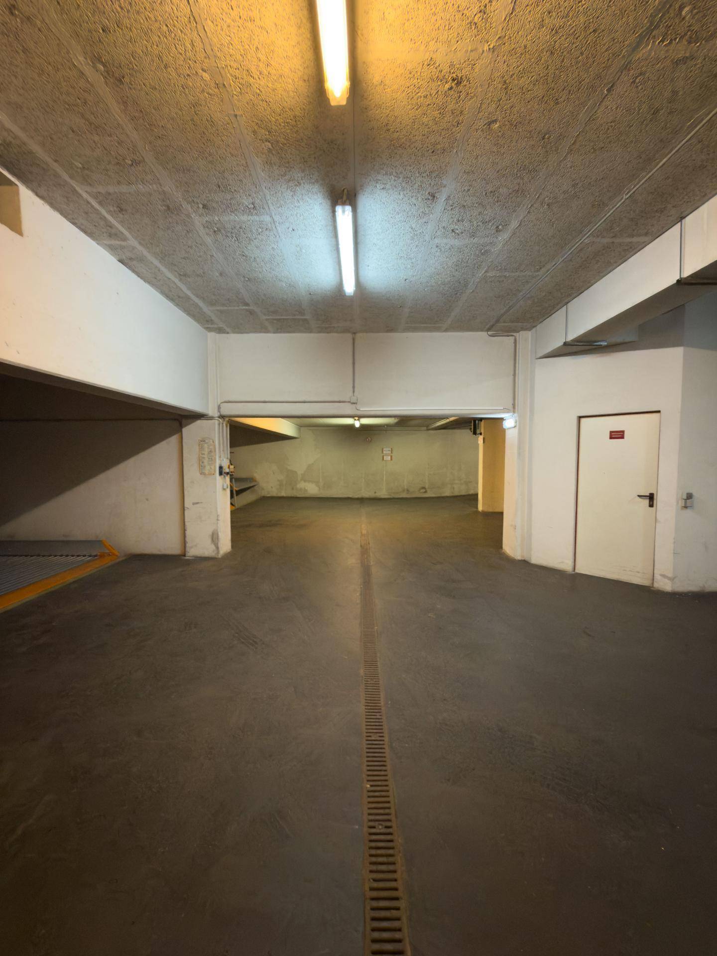 Geräumige Tiefgarage mit Betonwänden und -decke, ausgestattet mit Leuchtstoffröhren und einem Entwässerungssystem.