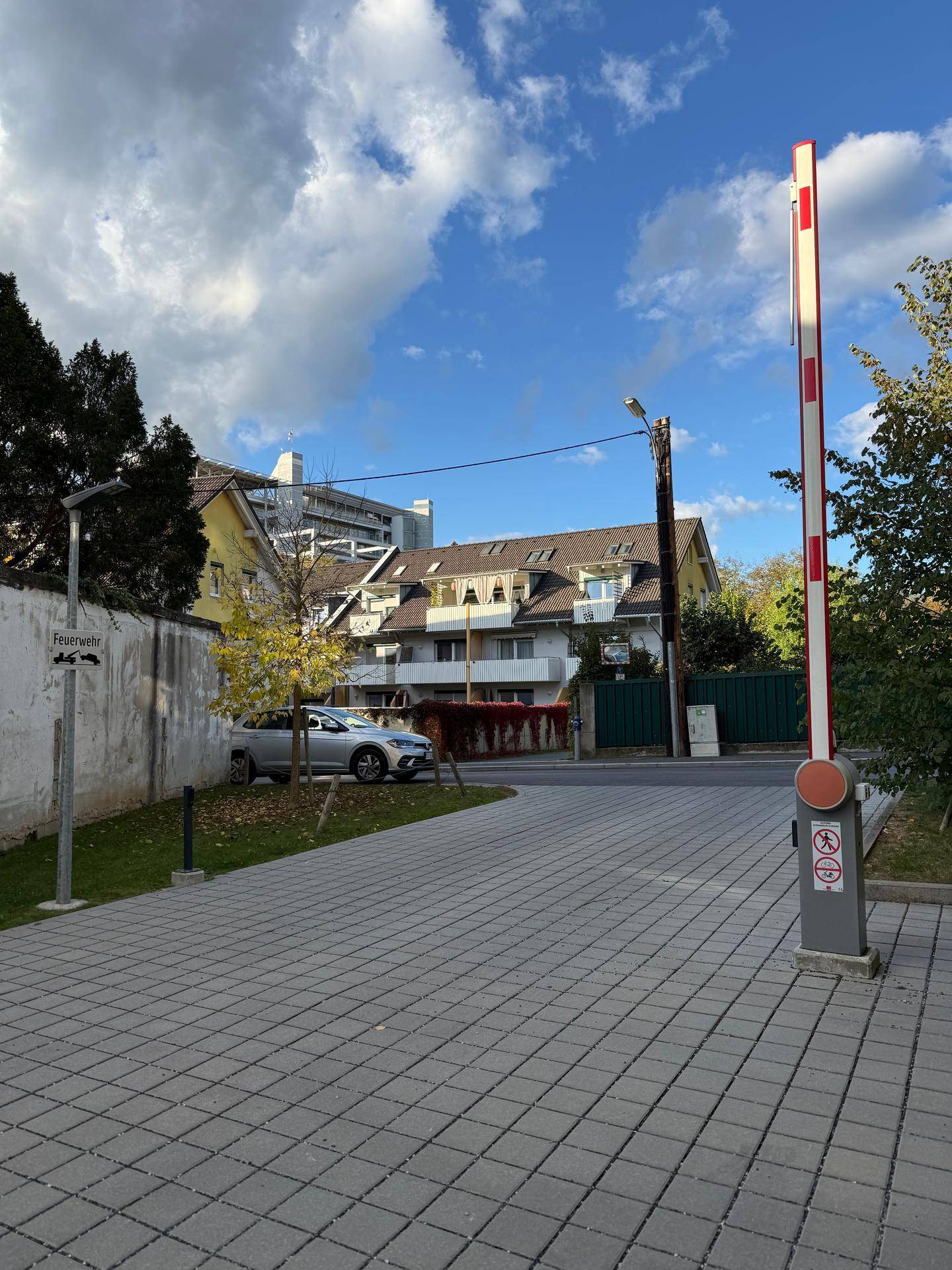 Zufahrt zu einem Wohngebäude mit Schranke, gepflastertem Bereich und parkenden Autos unter blauem Himmel.