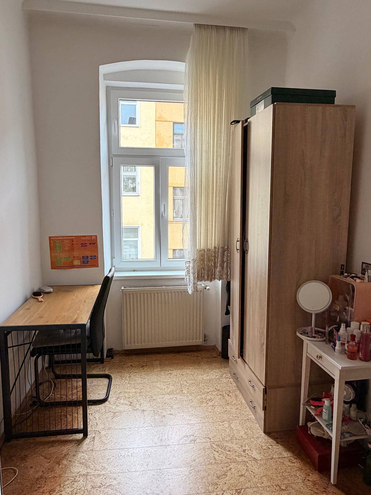 Helles Zimmer mit Schreibtisch, Kleiderschrank und Fenster mit Blick auf die Nachbargebäude.