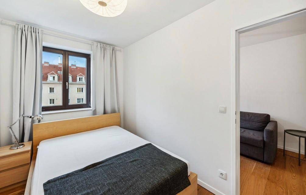 Helles Schlafzimmer mit Holzbett, Nachttisch und Fenster mit Blick auf die Dächer der Stadt.