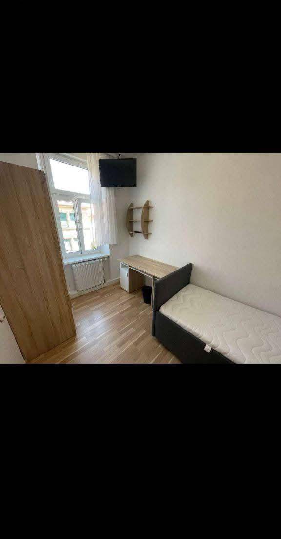 Schlafzimmer mit Fenster, Holzboden, einem Bett und einem an der Wand montierten Fernseher.