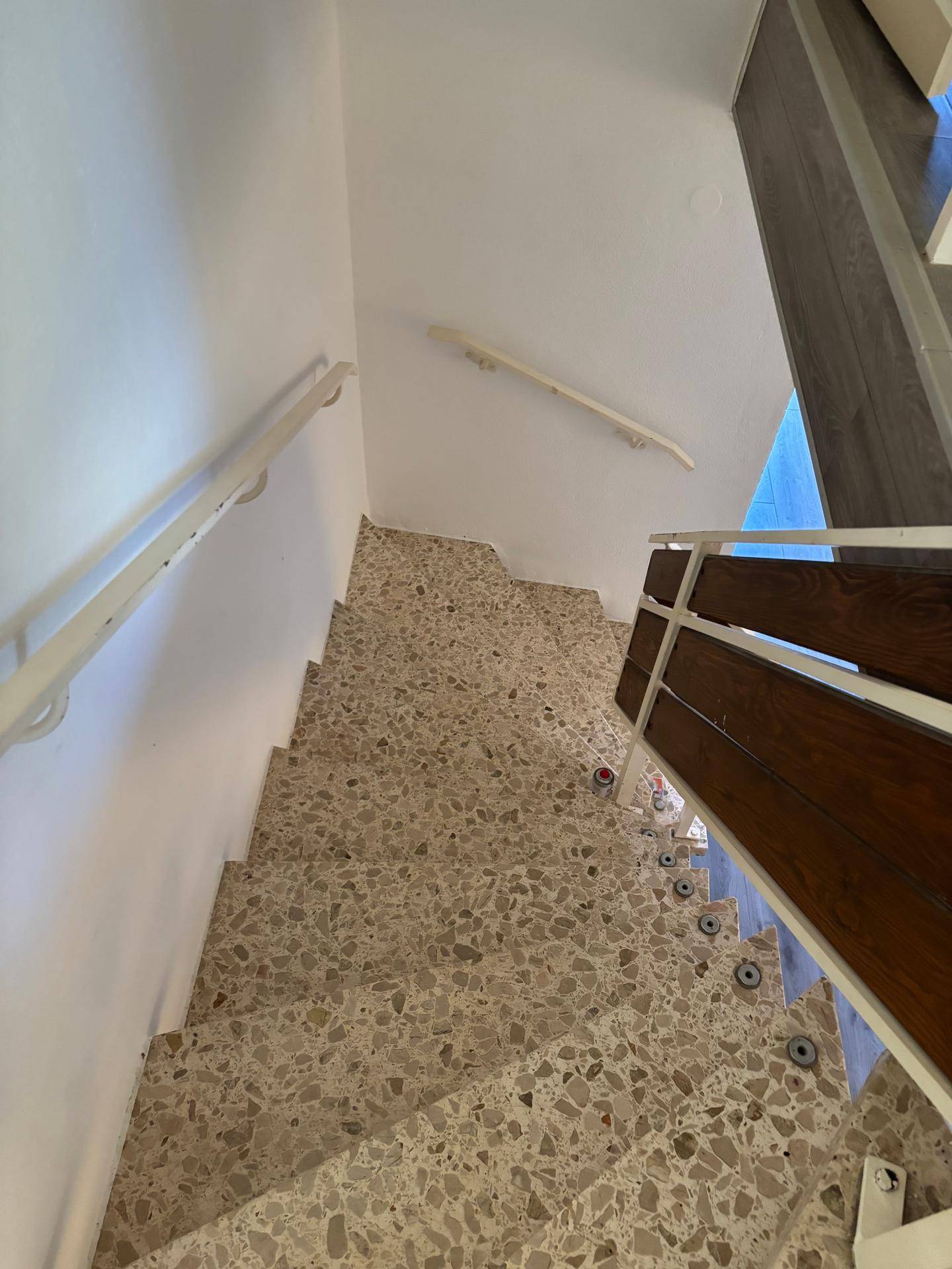 Treppenhaus mit Terrazzo-Boden, weiß gestrichenen Wänden und einem Holzgeländer.