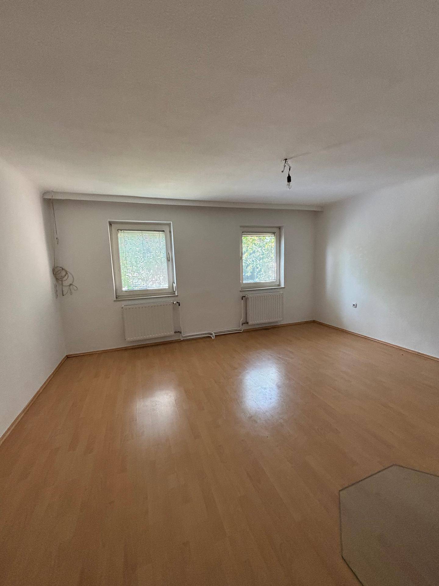 Helles, leeres Zimmer mit Laminatboden, weißen Wänden und zwei Fenstern mit Heizkörpern.