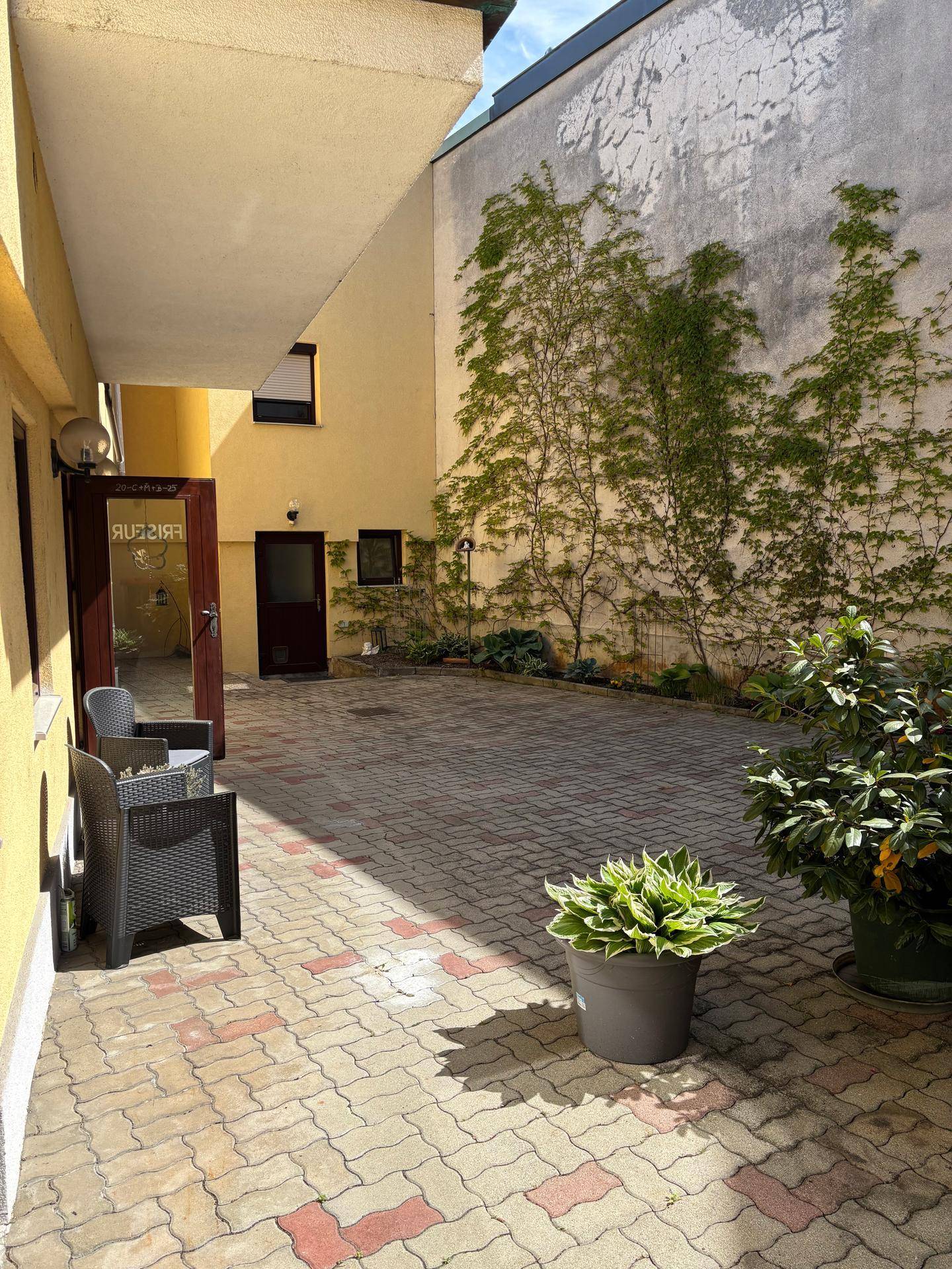Ein sonniger Innenhof mit Sitzgelegenheiten und einem traditionellen Holzbalkon am gelben Gebäude.