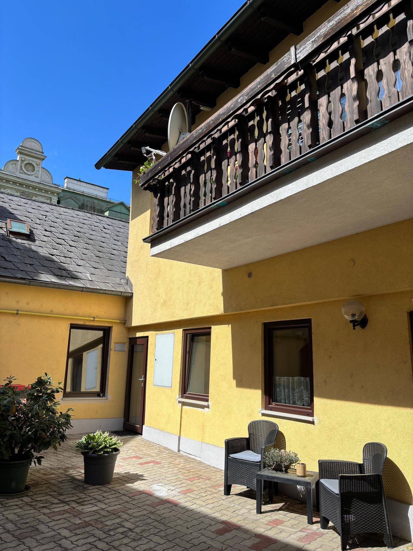 Ein sonniger Innenhof mit Sitzgelegenheiten und einem traditionellen Holzbalkon am gelben Gebäude.