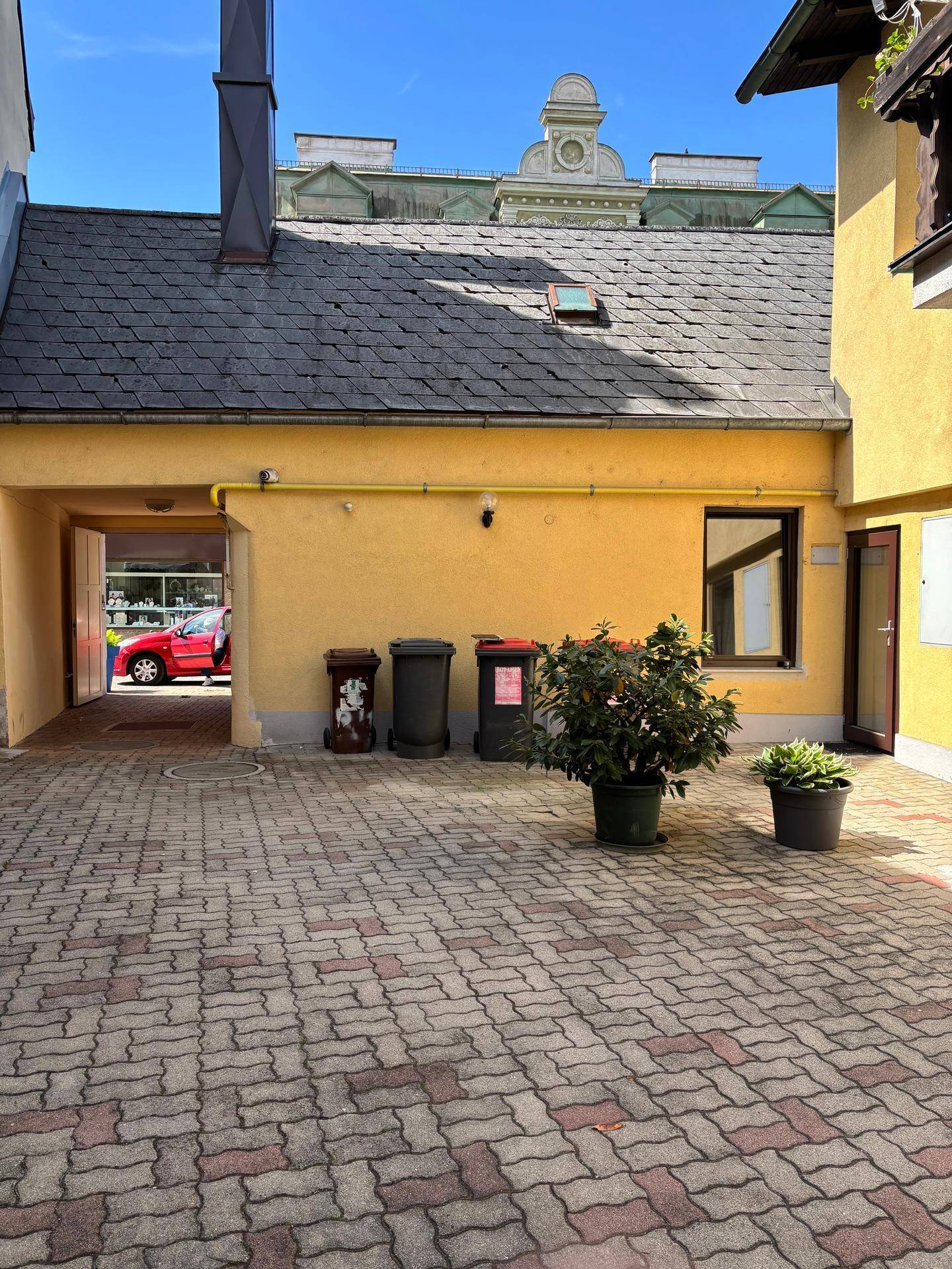 Ein gepflasterter Innenhof mit Mülltonnen und Pflanzen, der zu einem Durchgang führt.