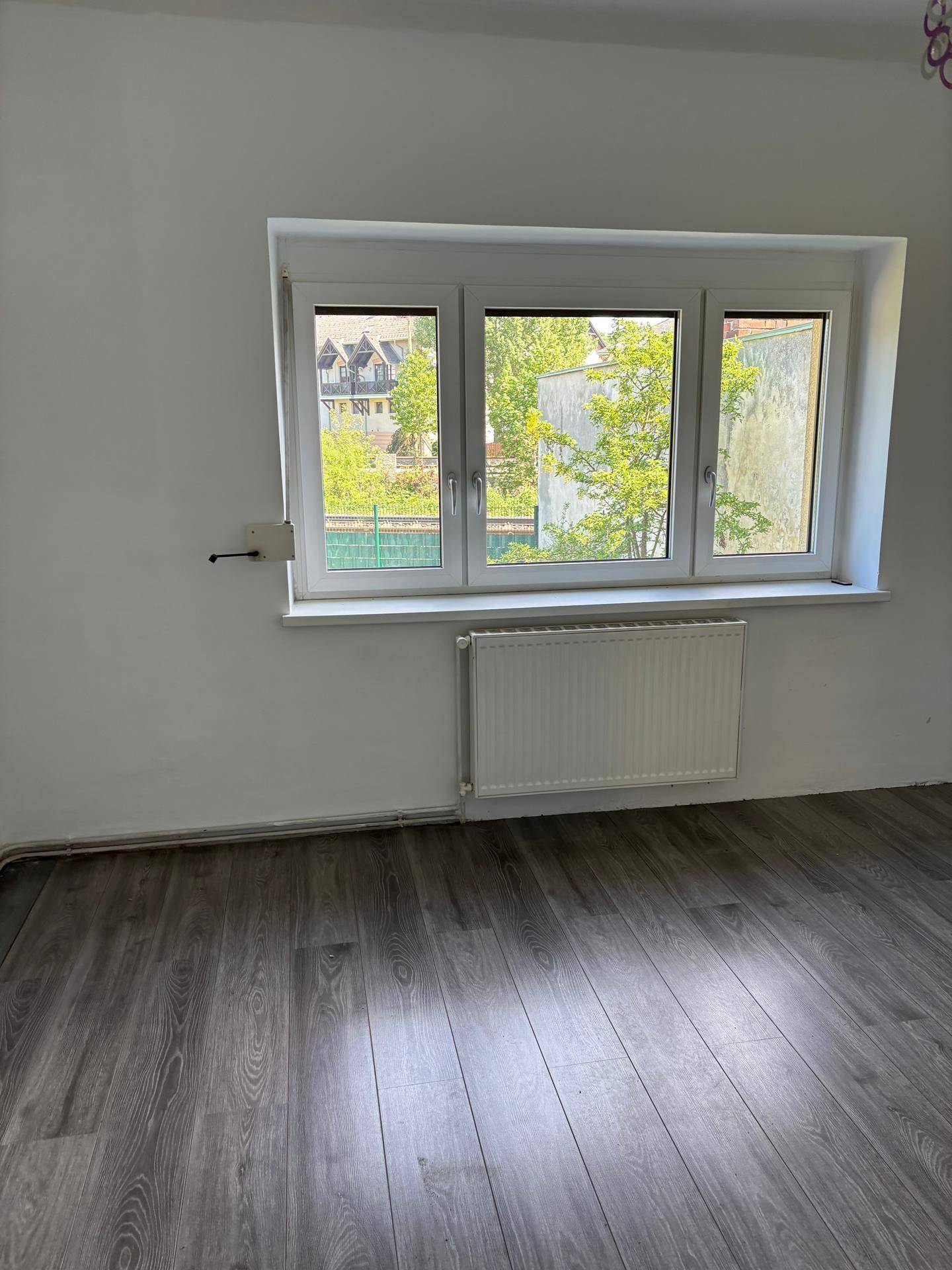 Ein helles Schlafzimmer mit grauem Laminatboden, weißen Wänden und einem großen Fenster.