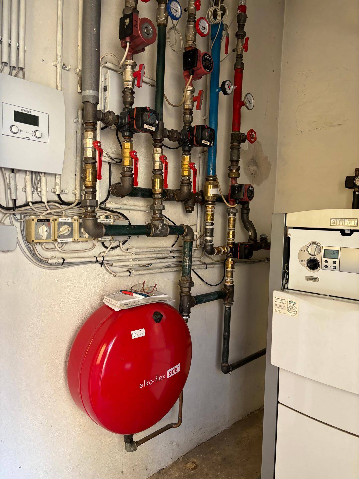Heizungsraum mit umfangreicher Rohrinstallation, einem roten Ausdehnungsgefäß und Steuerungselementen.