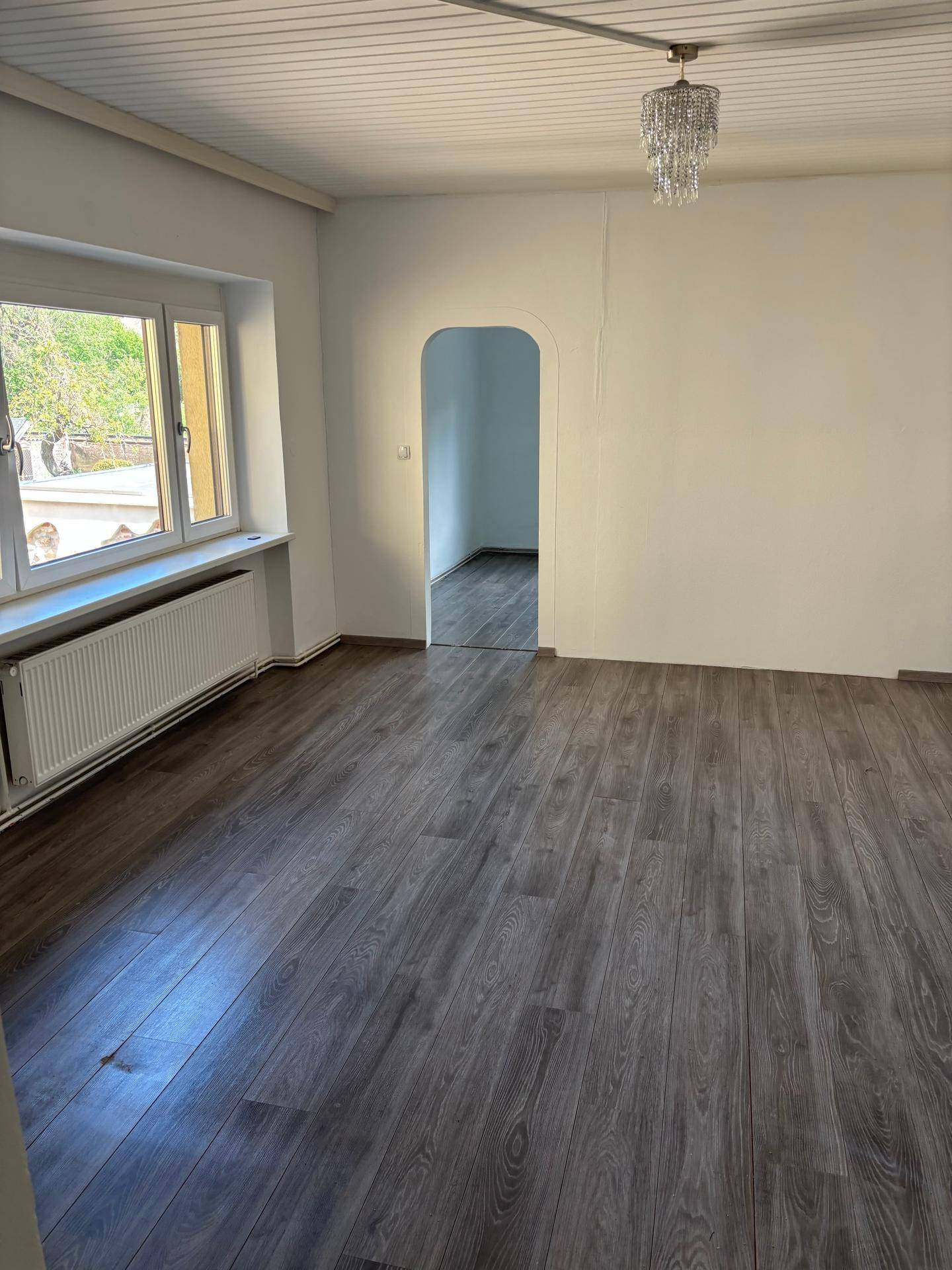 Ein heller Raum mit grauem Laminatboden, weißen Wänden und einem Fenster mit Heizkörper.