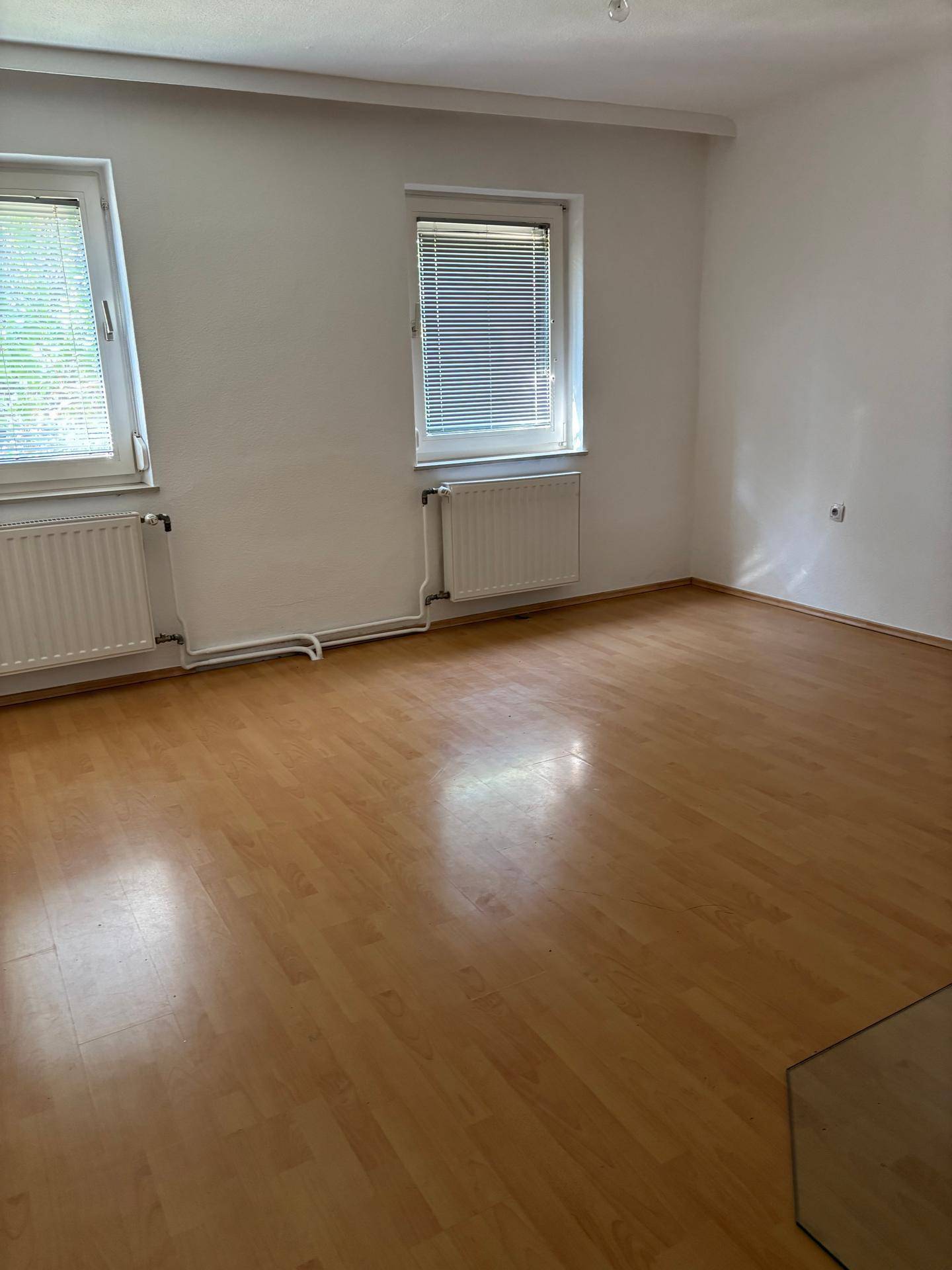 Helles, leeres Zimmer mit Laminatboden, weißen Wänden und zwei Fenstern mit Jalousien.