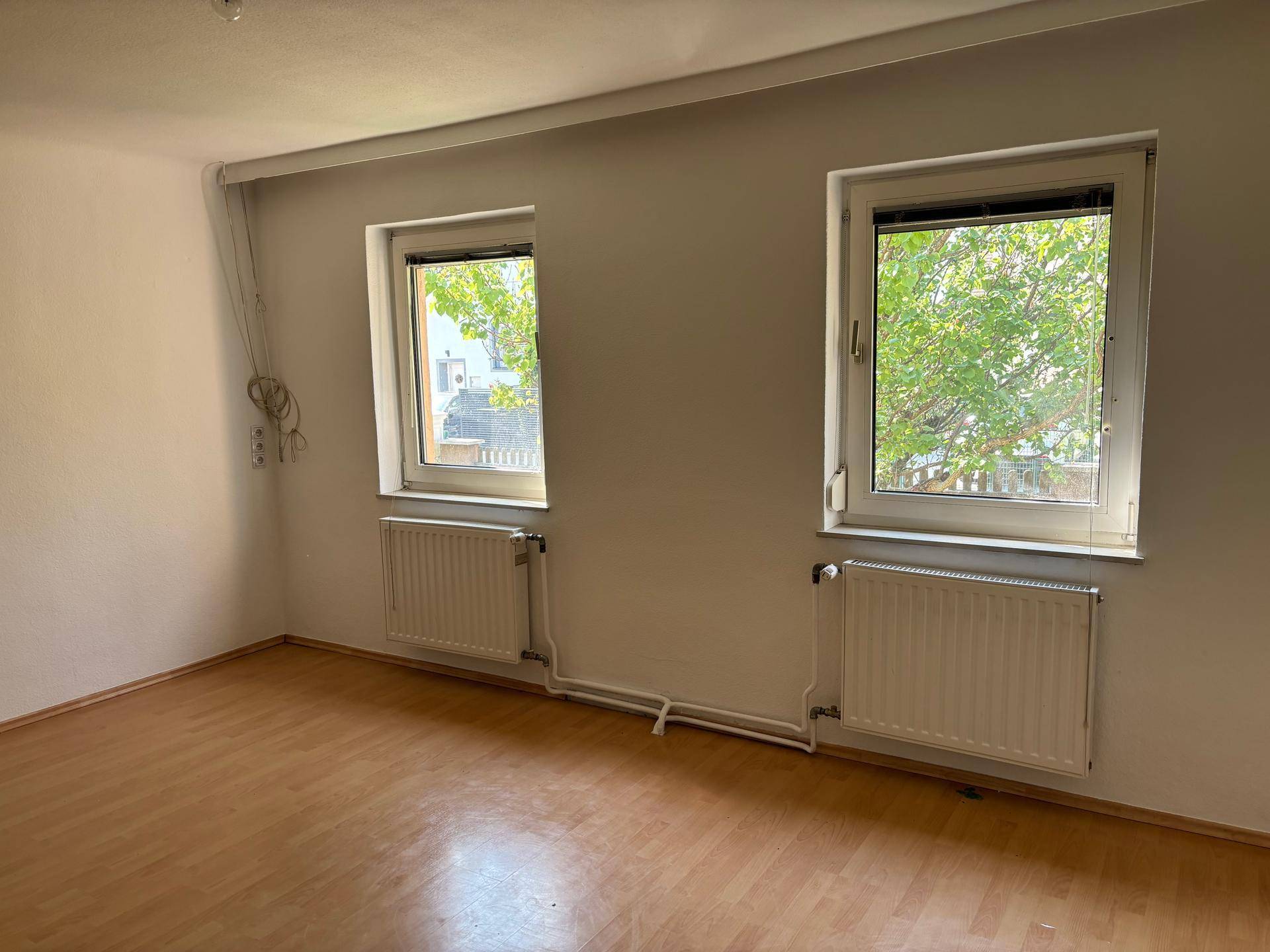Geräumiges Zimmer mit Laminatboden, weißen Wänden und zwei Fenstern, die eine grüne Aussicht bieten.