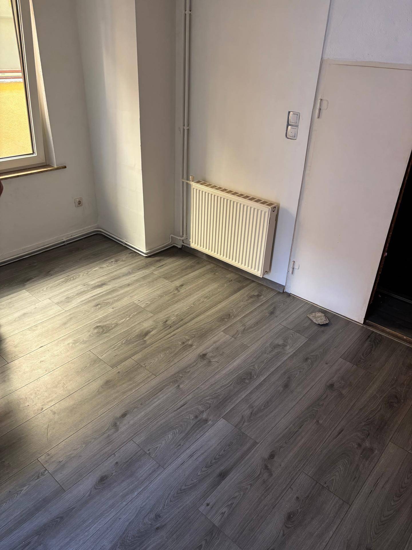 Leerer Raum mit Laminatboden, einem Fenster und einem Heizkörper an der Wand.