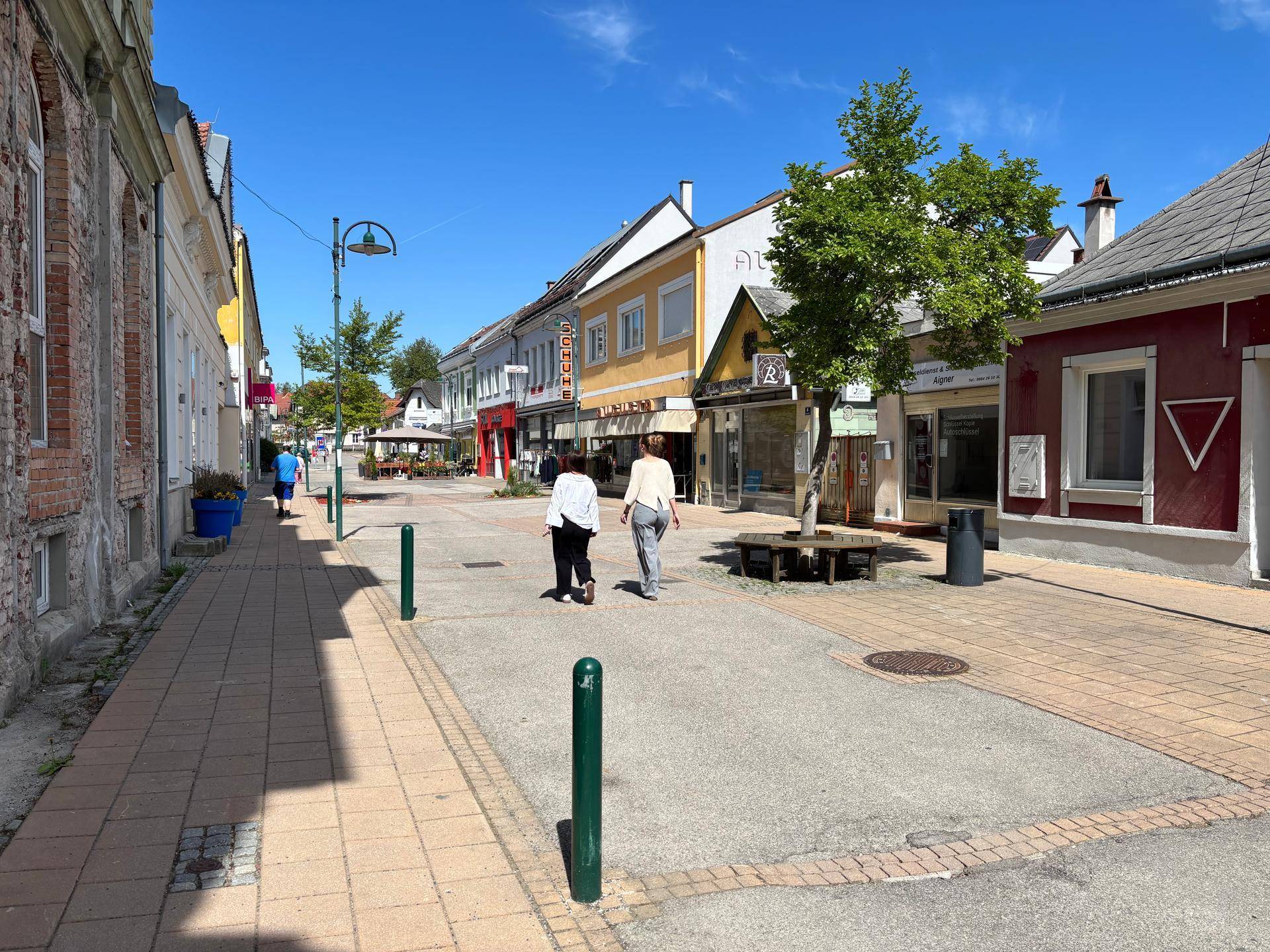 Eine belebte Einkaufsstraße mit verschiedenen Geschäften und Fußgängern an einem sonnigen Tag.
