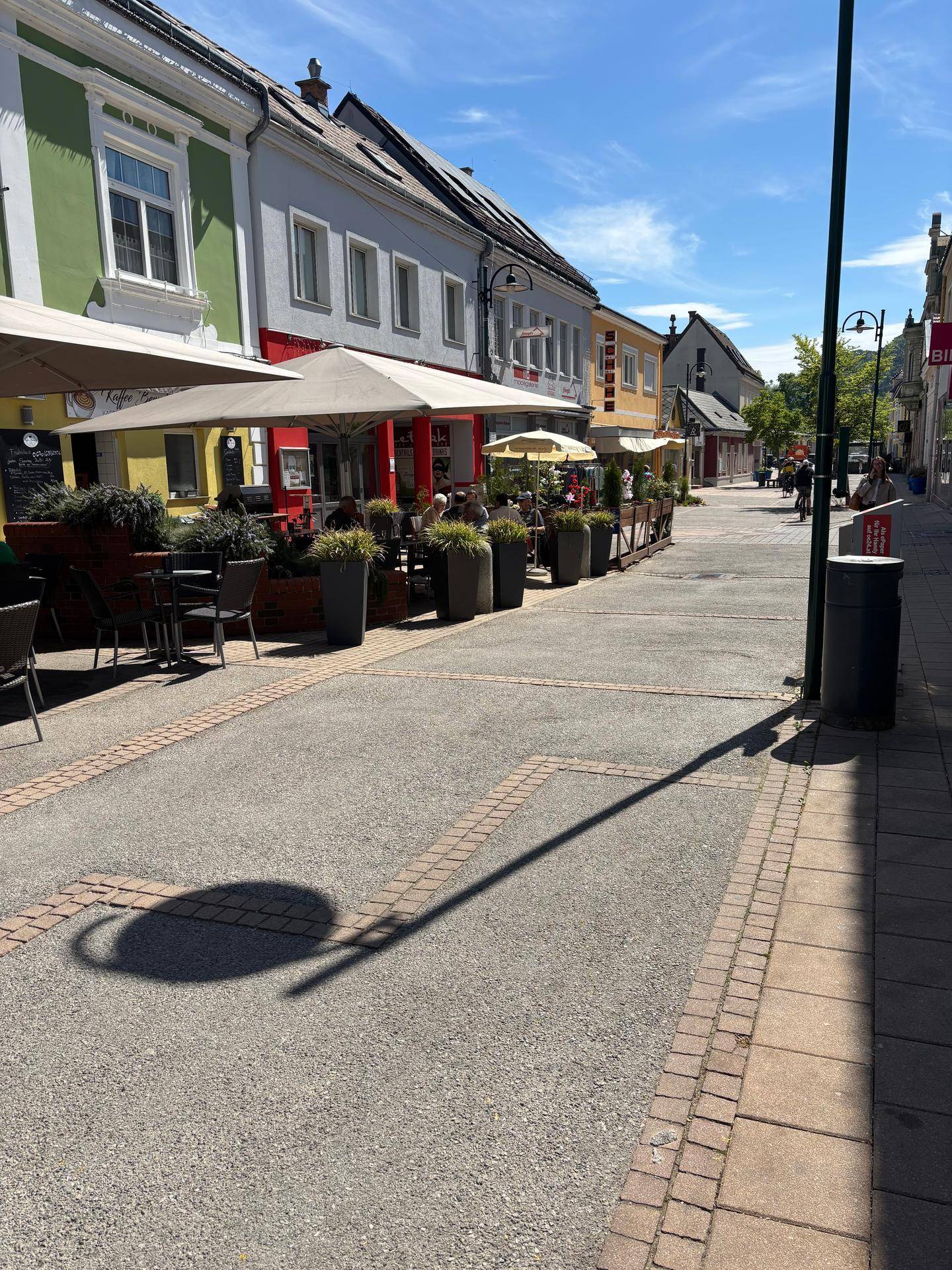 Belebte Einkaufsstraße mit Cafés, Geschäften und Fußgängern unter strahlend blauem Himmel.