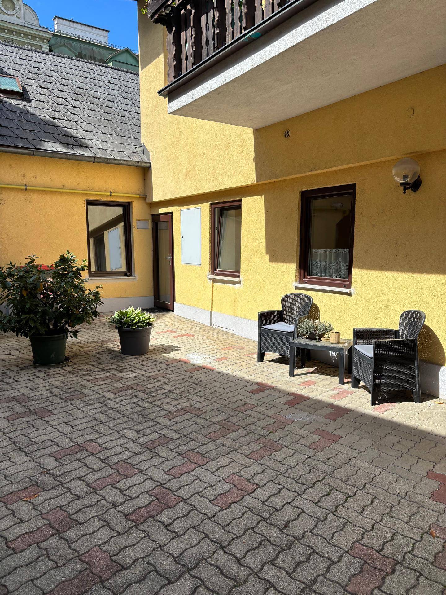 Ein gepflasterter Innenhof mit Sitzgelegenheiten und Topfpflanzen an einer gelben Fassade.