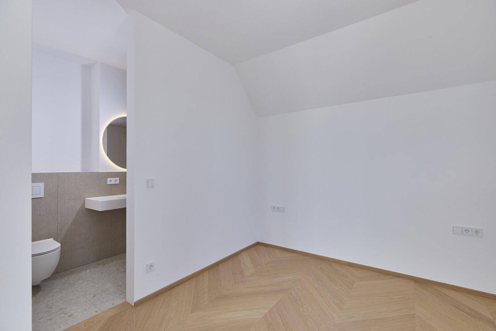 Schlafzimmer mit Fischgrätparkett und direktem Zugang zu einem modernen Badezimmer.