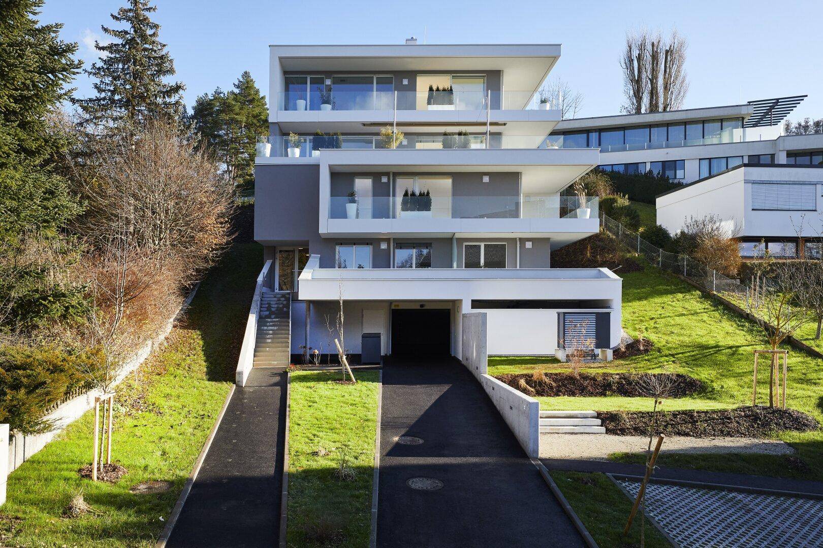 Modernes Mehrfamilienhaus mit gestuften Balkonen, großen Fenstern und Tiefgaragenzufahrt an einem Hang.