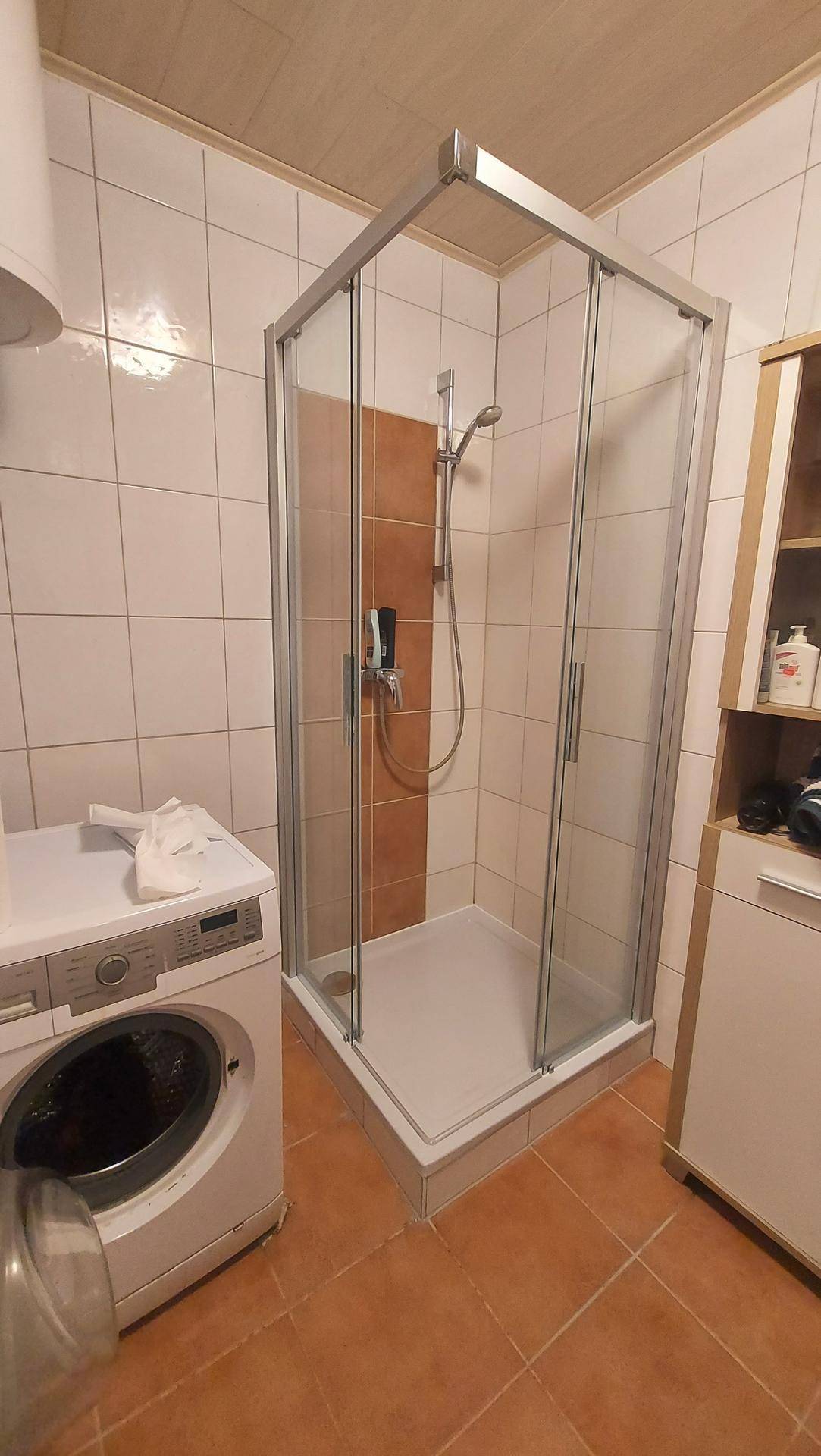 Badezimmer mit Duschkabine, Waschmaschine und hellen Fliesen an Wänden und Boden.