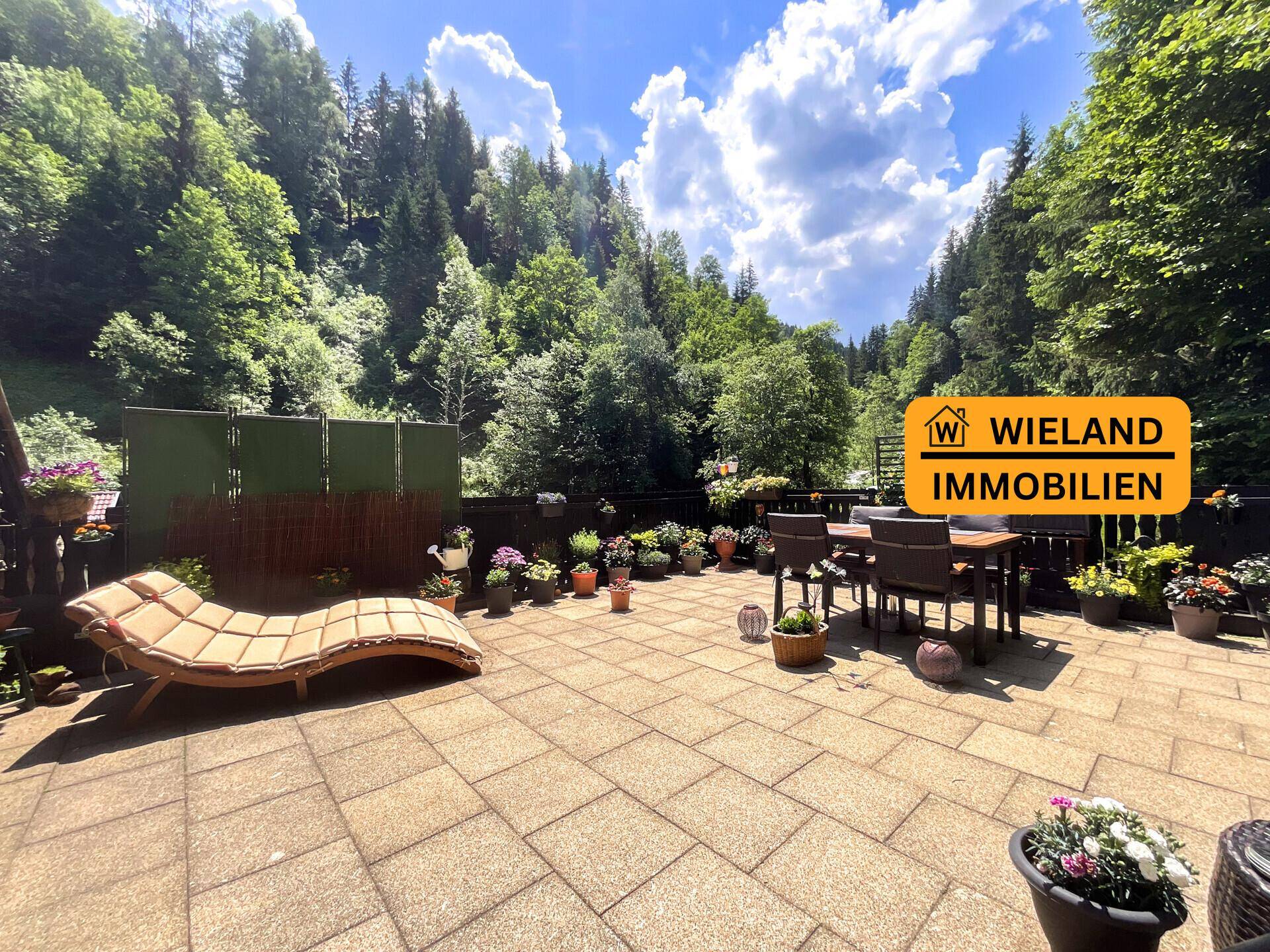 Große gepflasterte Terrasse mit Gartenmöbeln und Liegefläche, umgeben von bewaldeten Hügeln.