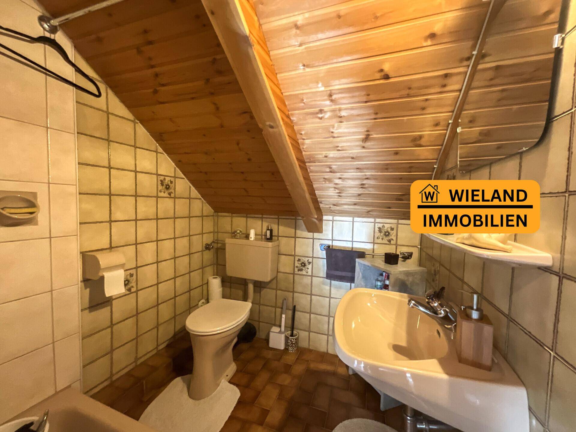 Badezimmer mit schrägen Holzwänden, beigefarbenen Fliesen und einfacher Ausstattung, inklusive Toilette und Waschbecken.