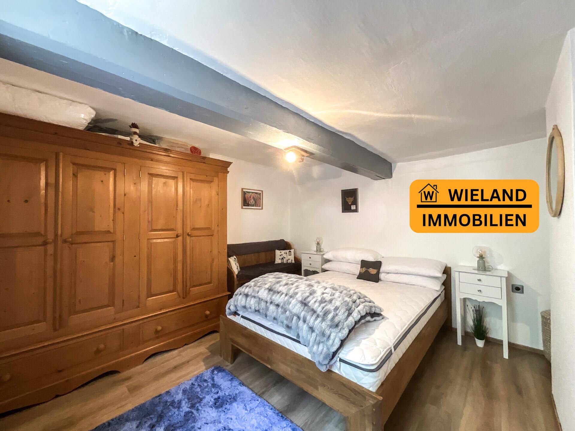 Schlafzimmer mit weiß gestrichenen Wänden, dunklen Deckenbalken und großem Holzschrank, bietet viel Stauraum.