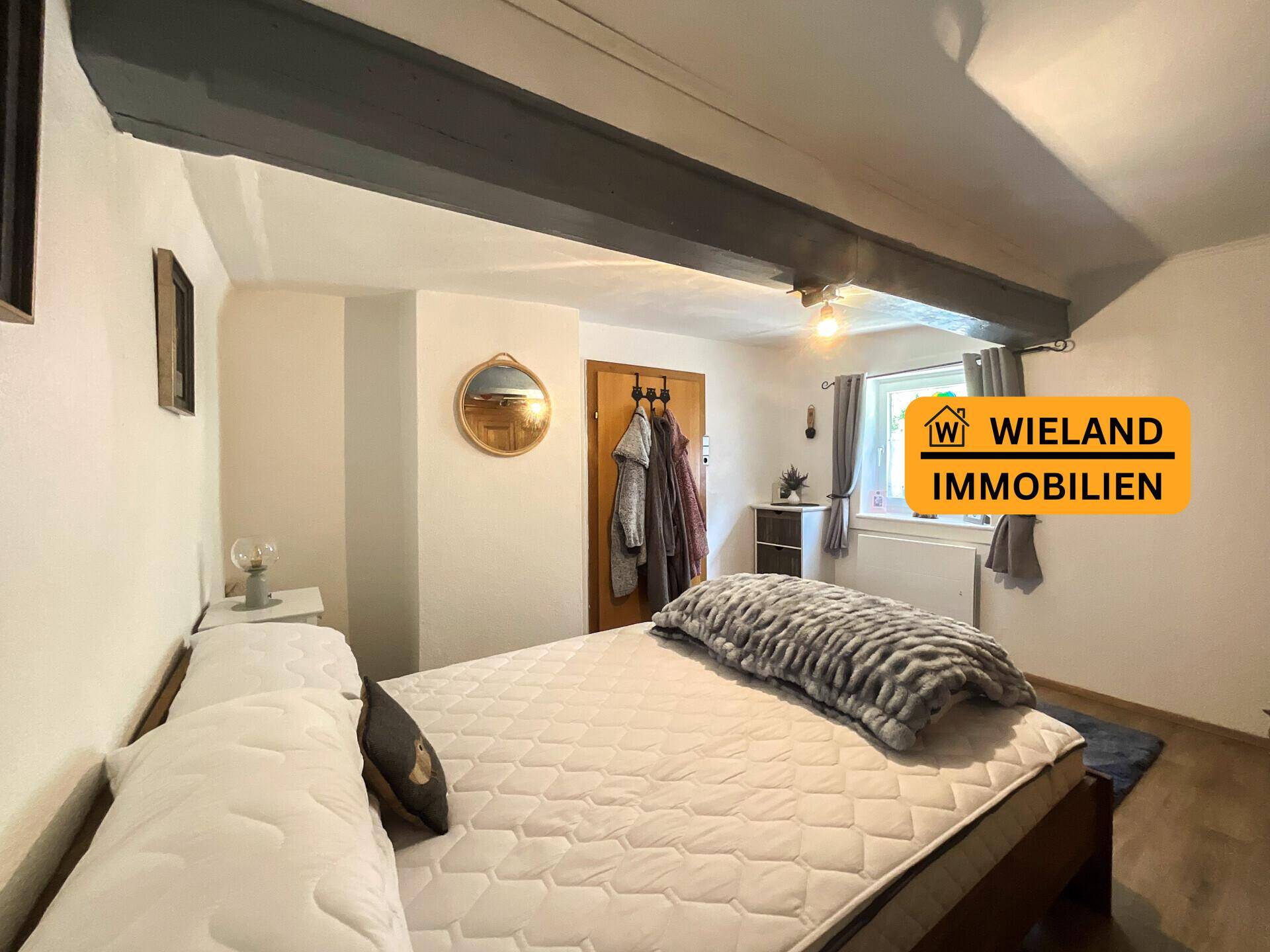 Helles Schlafzimmer mit sichtbaren Deckenbalken, Doppelbett und Fenster, das viel Tageslicht hereinlässt.