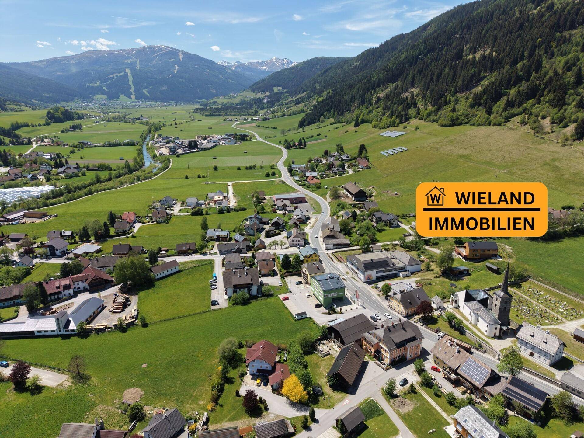 Weitläufige Luftaufnahme des Dorfes in einem Tal, umgeben von grünen Wiesen und Bergen.