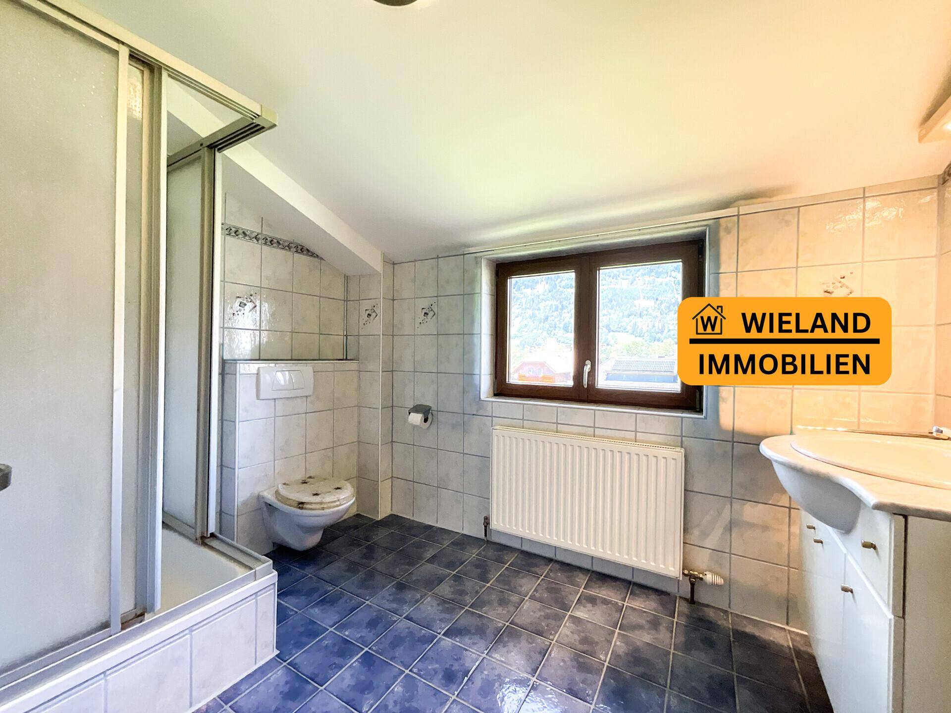 Helles Badezimmer mit blauen Bodenfliesen, Duschkabine, WC und Fenster.