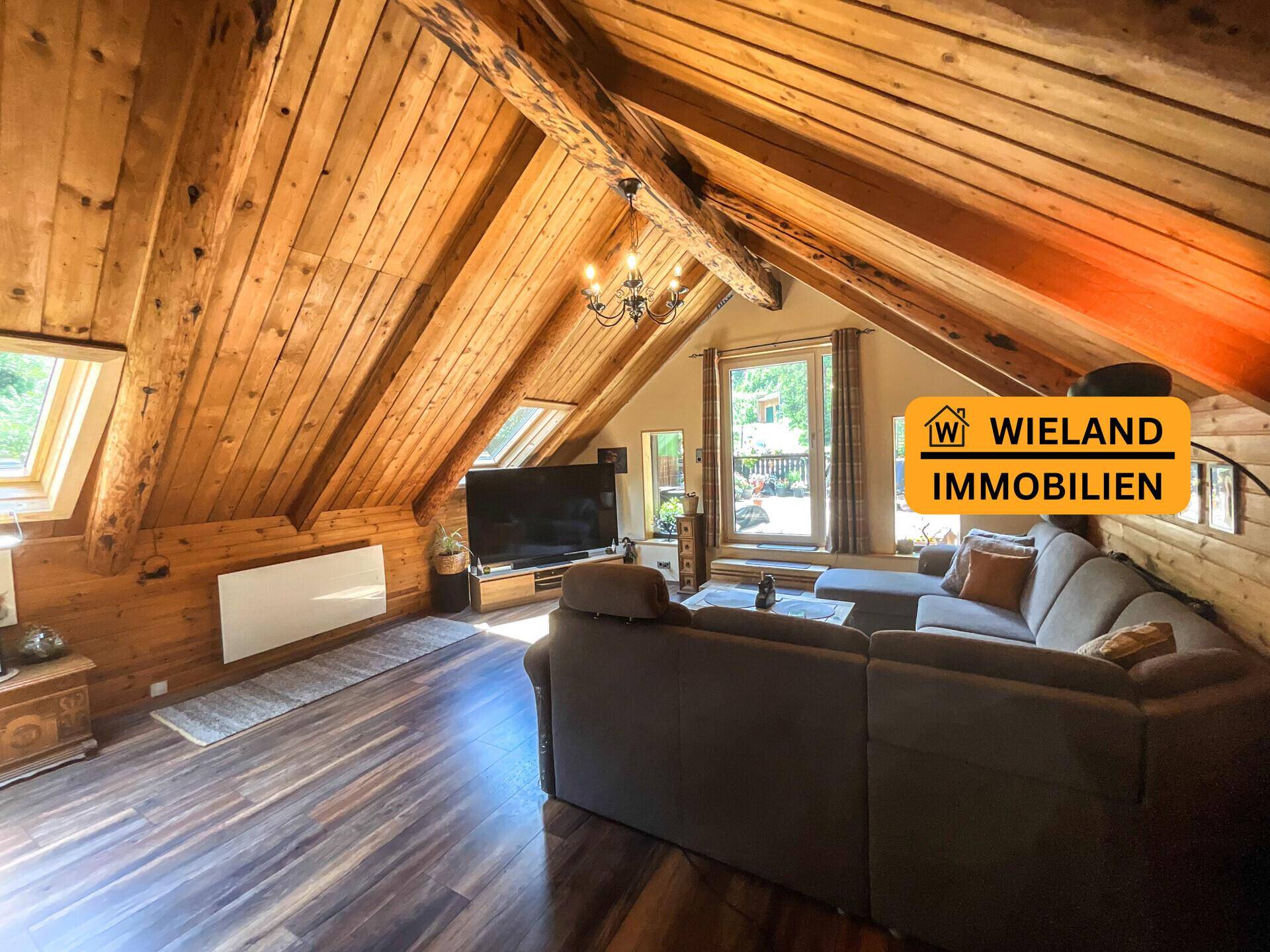 Wohnbereich unter dem Dach mit Holzbalken, gemütlichem Sofa und Blick ins Grüne durch die Fenster.