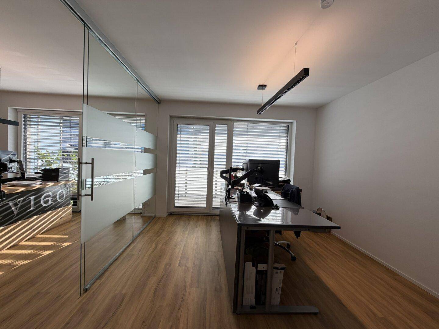 Modernes Büro mit Glaswand, großen Fenstern und Jalousien für angenehmes Arbeitslicht.