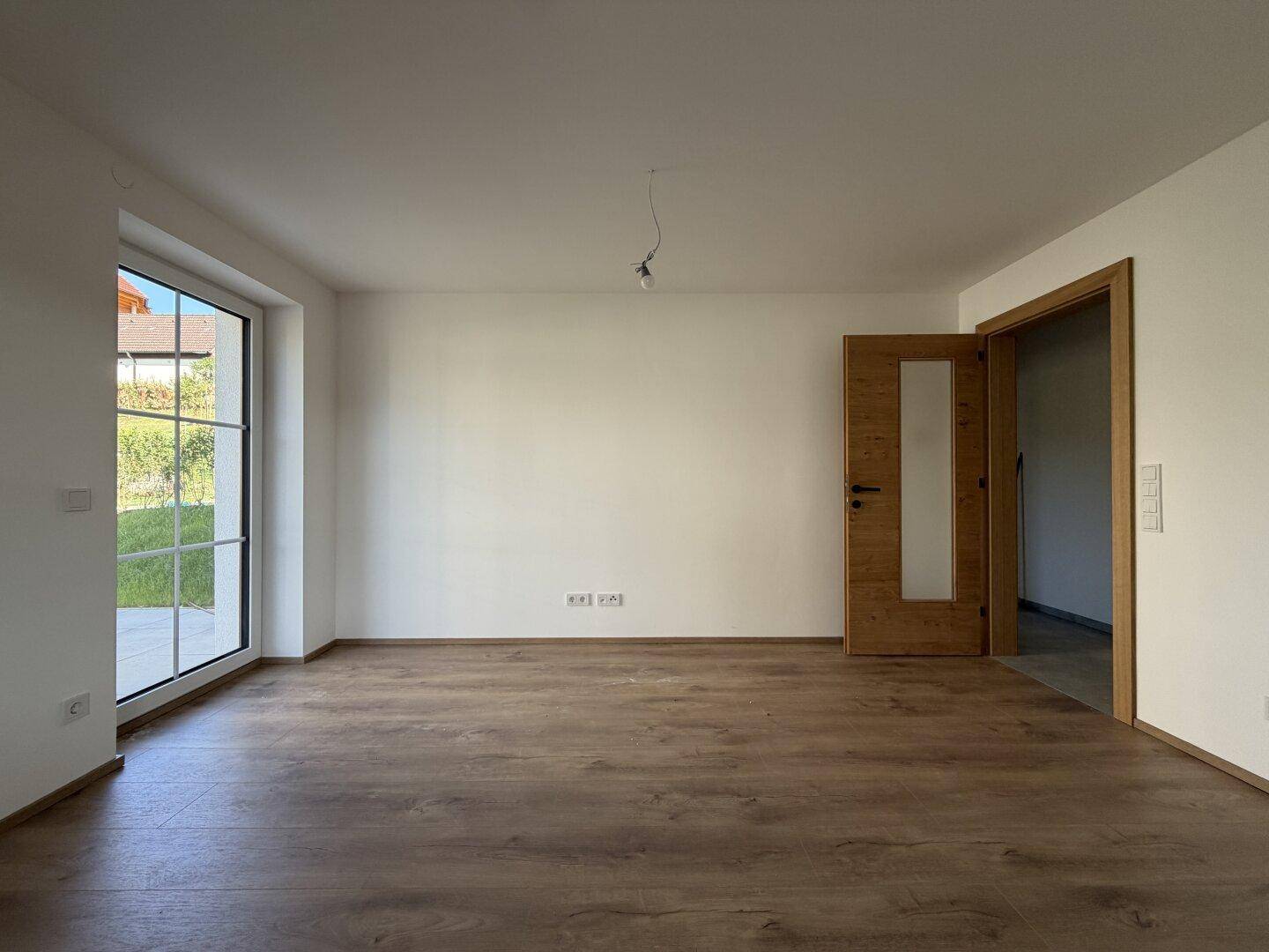 Heller, leerer Raum mit Laminatboden und großem Fenster mit Blick ins Grüne.