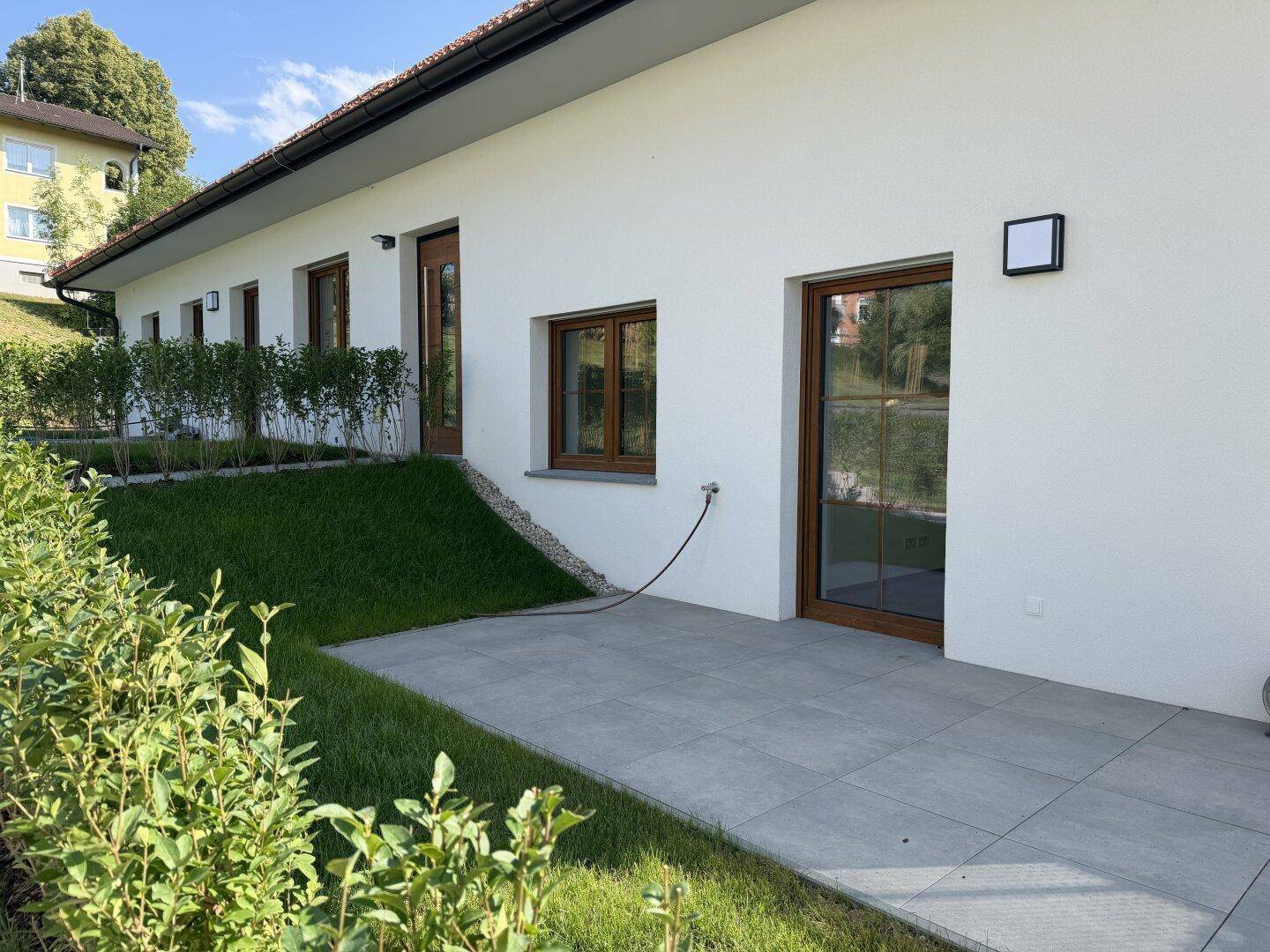 Seitenansicht des Hauses mit einer kleinen, sonnigen Terrasse und grüner Bepflanzung.