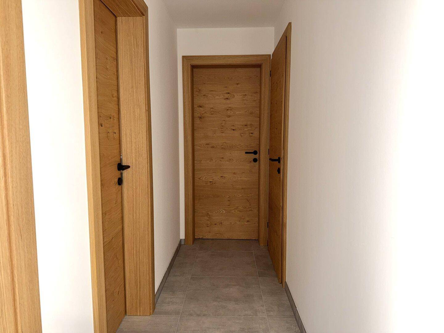 Heller Flur mit grauen Fliesen und mehreren Holztüren, die zu den Zimmern führen.