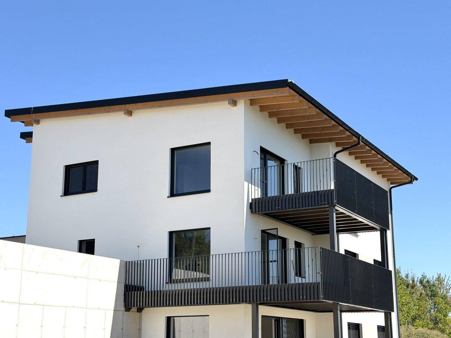 Modernes Wohngebäude mit weißer Fassade, schwarzen Balkonen und Holzdachkonstruktion.